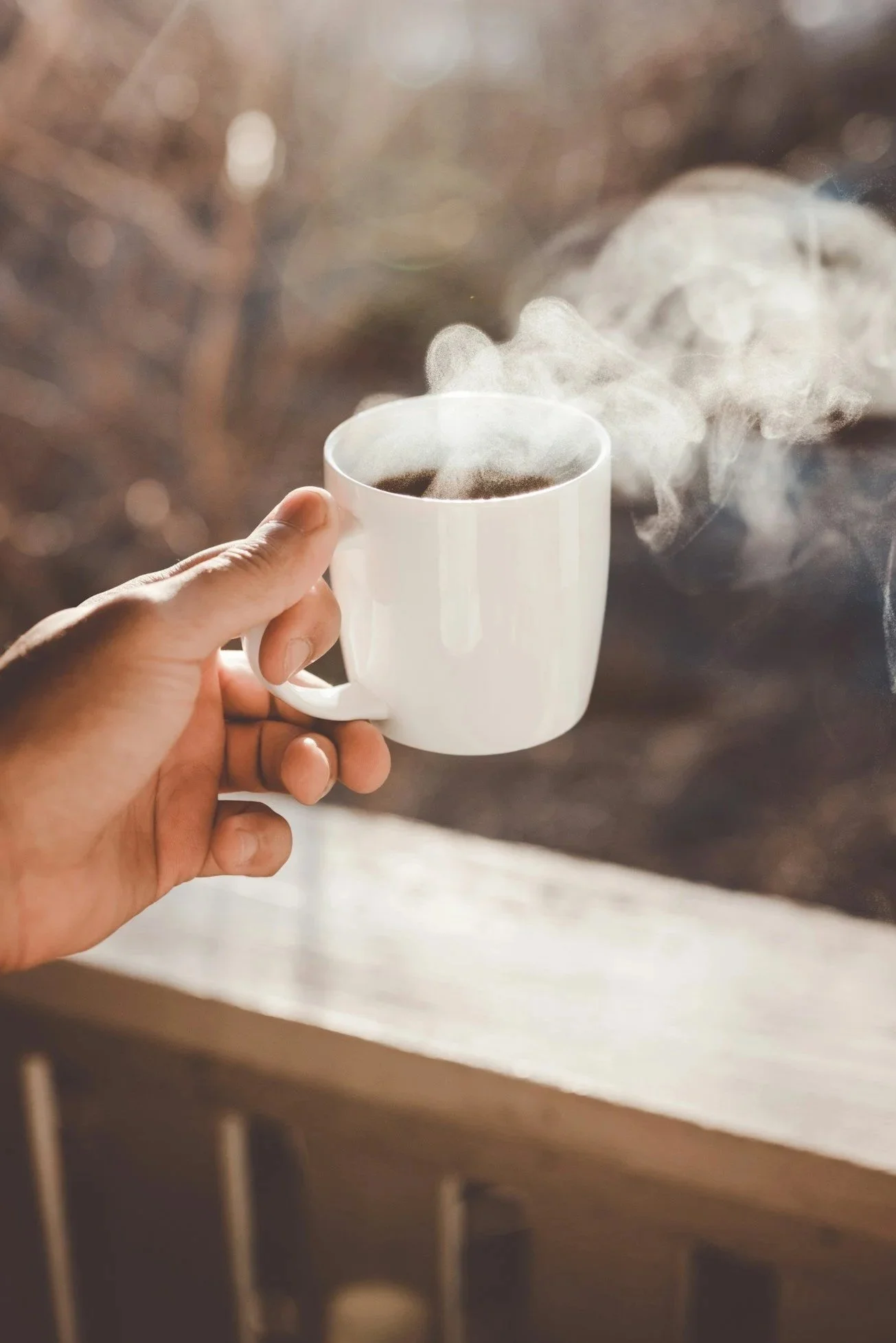A hand holding a white mug filled with hot coffee, with steam rising from it, against a blurred background.