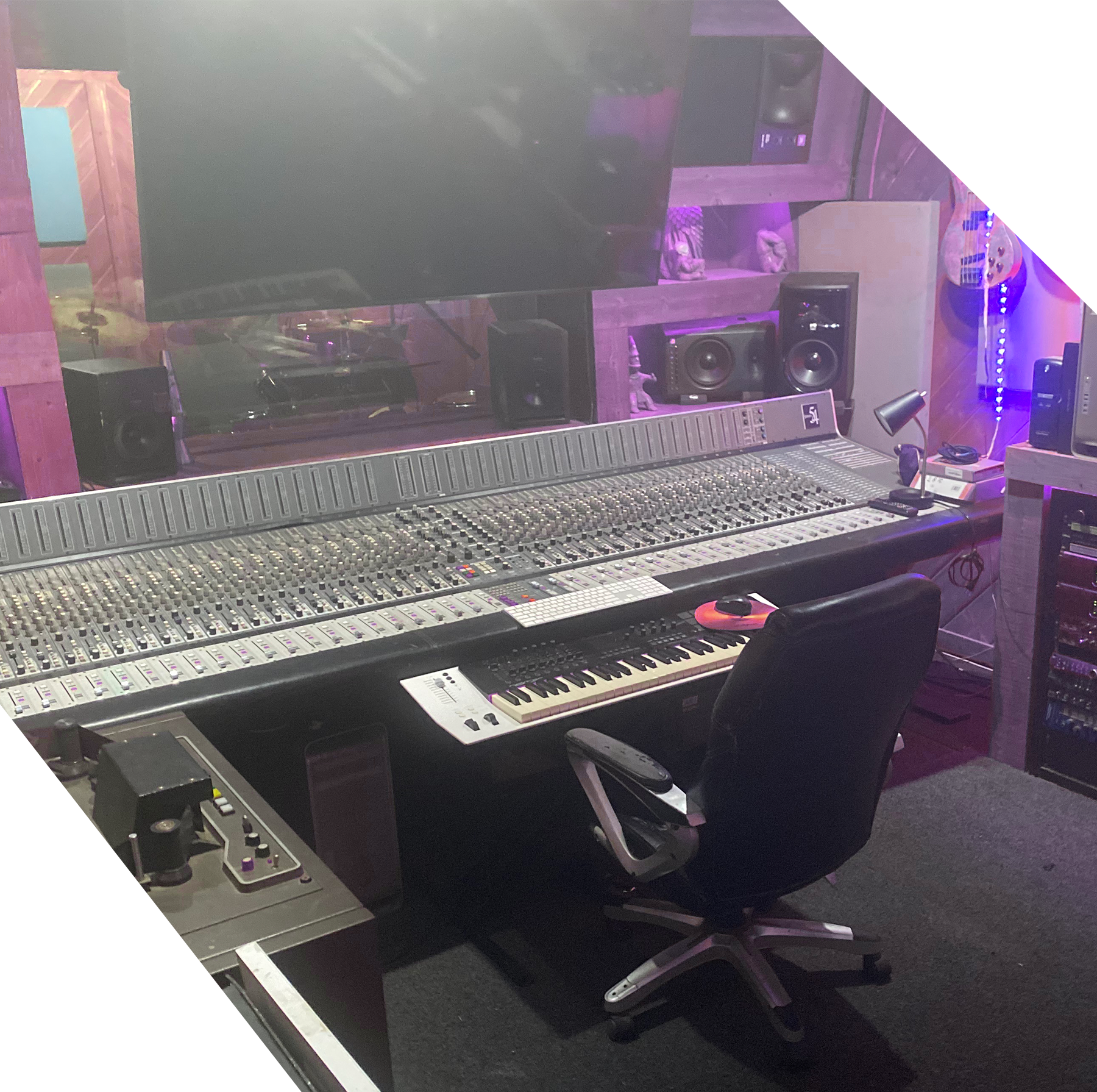 Music production studio with large mixing console, computer keyboard, keyboard synthesizer, studio monitor speakers, and acoustic panels with purple lighting.