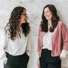 Two women with dark, curly hair and dark, straight hair smiling and chatting indoors.