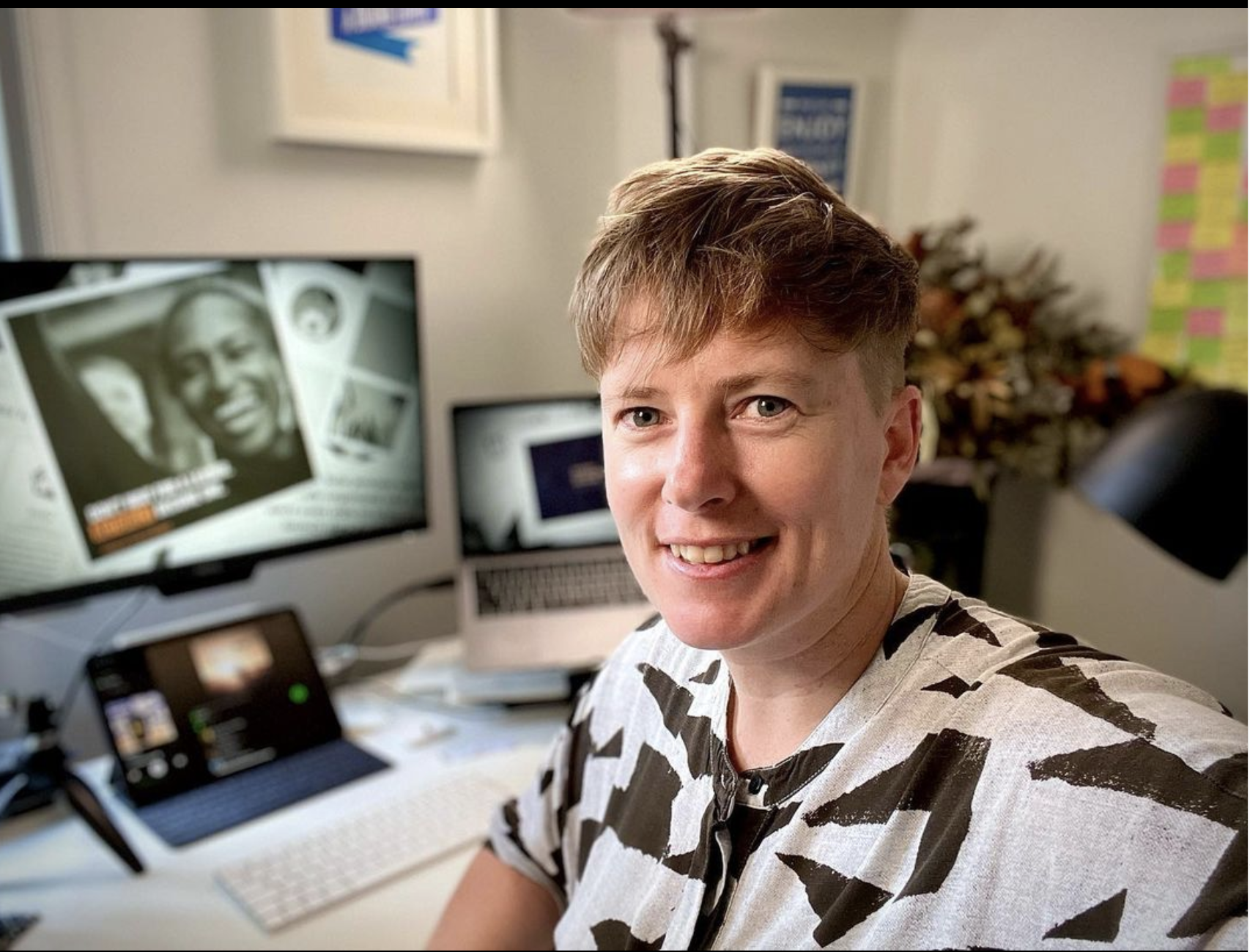 A smiling person with short, light brown hair in a white and black patterned shirt taking a selfie in a home office. Behind them are a desktop monitor displaying a magazine cover, a laptop, and a tablet on a stand. The background includes some wall decorations, a colorful sticky note wall, and a plant.