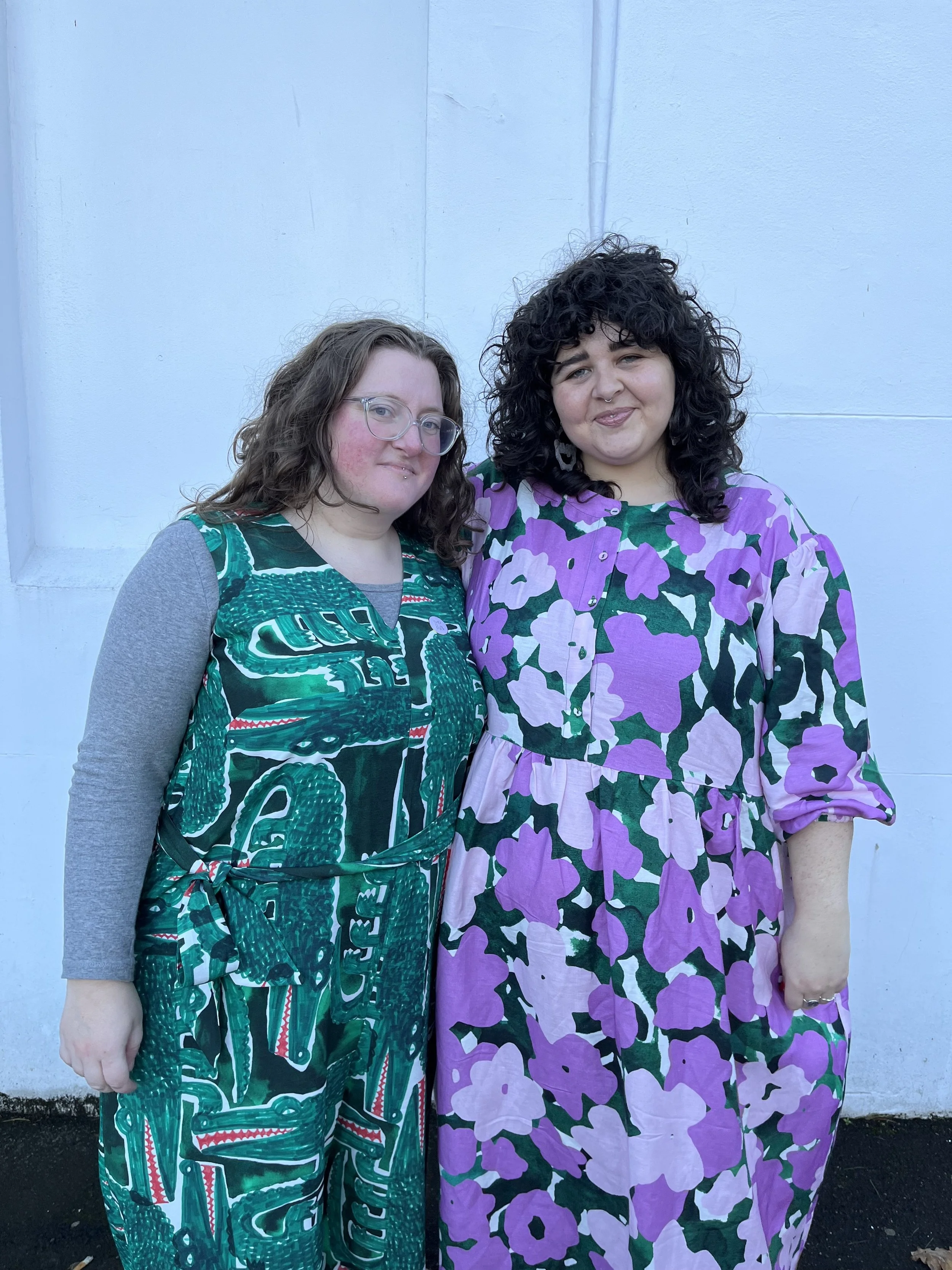 Two women standing close together in front of a white wall, smiling at the camera. One is wearing glasses and a jumpsuit with abstract green, black, and white patterns, and the other is wearing a purple floral dress with a rainbow pattern.