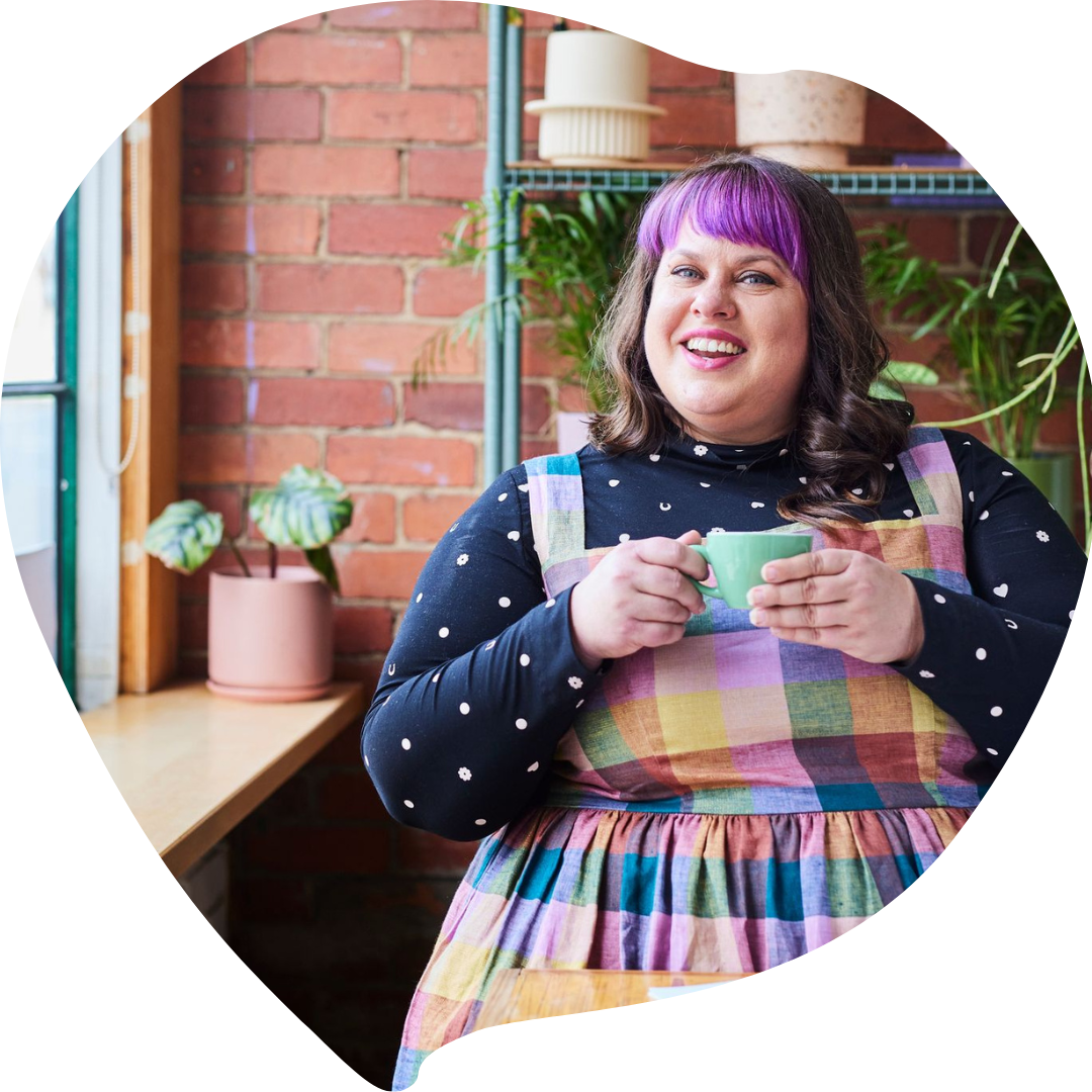 A smiling woman with purple hair and a colorful checkered apron, holding a green mug, standing indoors near a window with potted plants and a brick wall background.