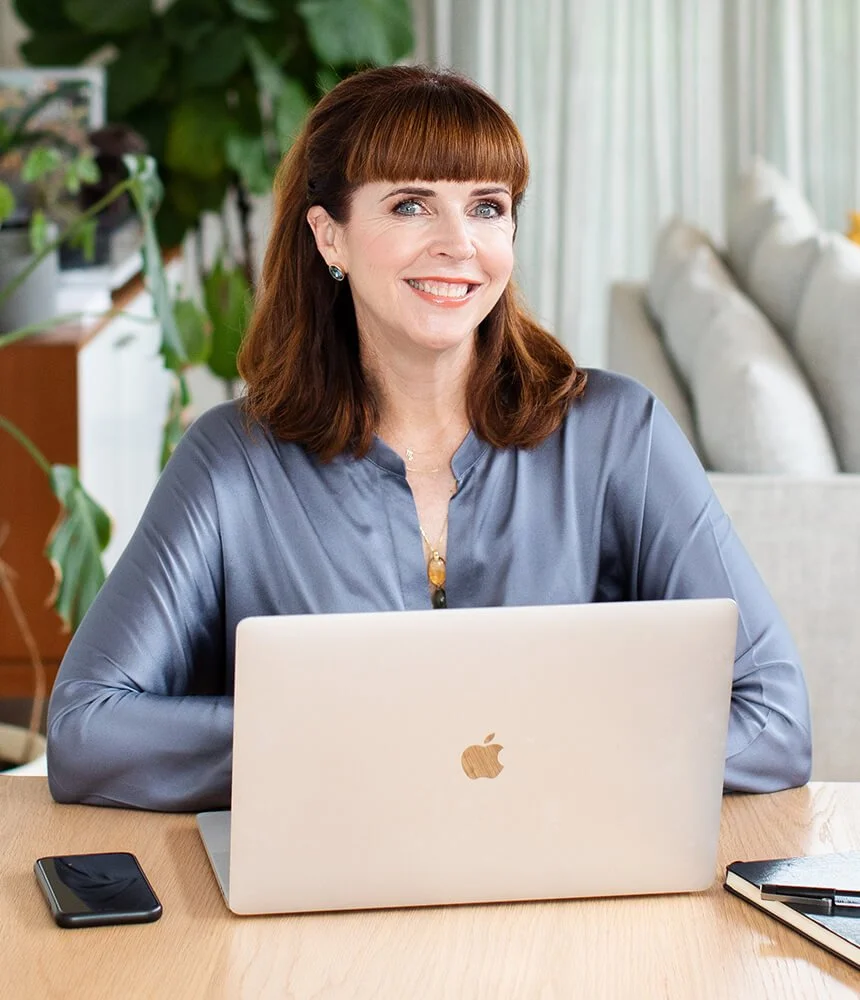 Smiling woman with red hair and bangs sitting at a desk with a silver MacBook, a smartphone, and a notebook, in a bright room with green plants and white curtains.