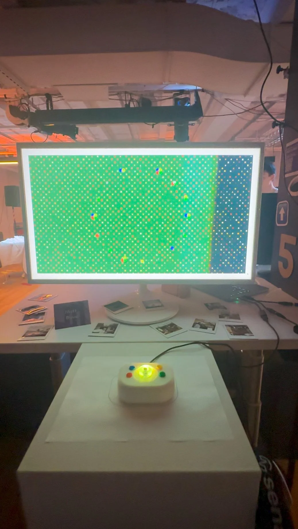 A computer monitor displaying a pixelated or glitched green background with colorful dots. In front of the monitor, there is a white game controller with colorful buttons and an indicator light. The setup is on a white table with printed photos and cards around it, and the scene appears to be at an indoor event or exhibition.