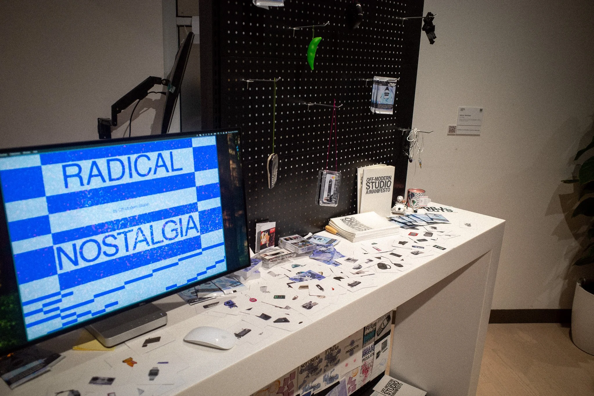 A display table with scattered small printed images, a computer monitor showing the word "RADICAL NOSTALGIA" in blue text on a pixelated background, and a black pegboard with hanging items including books, cards, and small objects in a gallery or exhibition space.