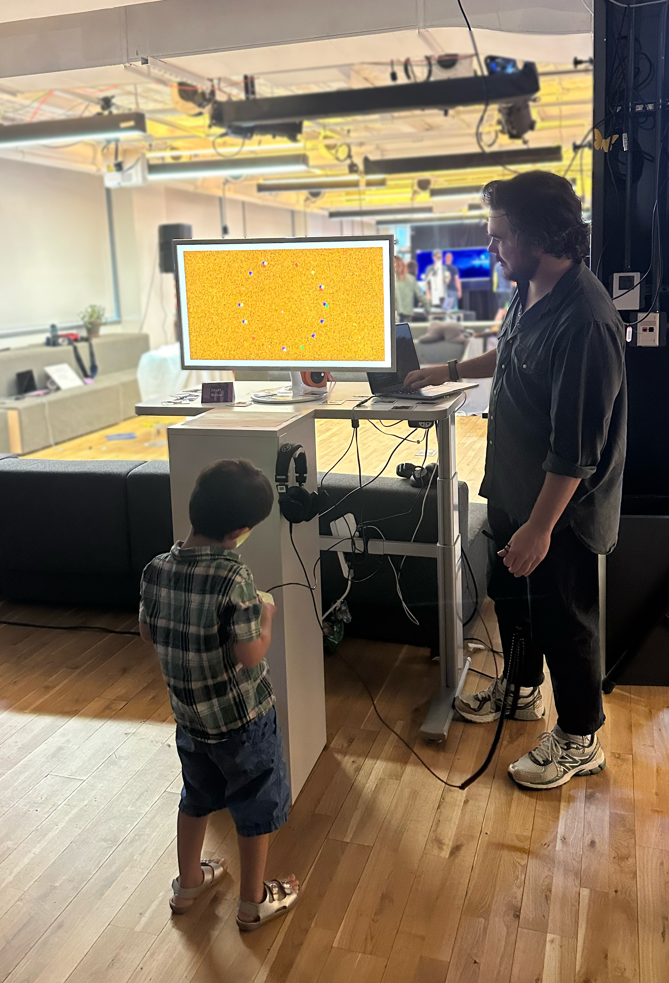 A young boy and a man with prosthetic leg are standing in front of a monitor in an indoor setting. The monitor displays a colorful game or activity with small objects or balls. The man is standing next to a small table with a laptop, and the boy is looking at the screen.