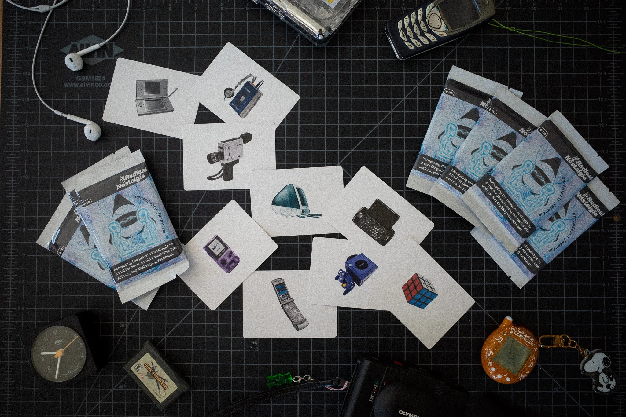 A black work surface with scattered electronic items, photo cards, and paper promotional packs of 'Radical Nostalgia' cards. Items include a pair of white earphones, a small clock, a vintage handheld game console, a Rubik's Cube, a remote, a walkie talkie, and a keychain with Snoopy attached.