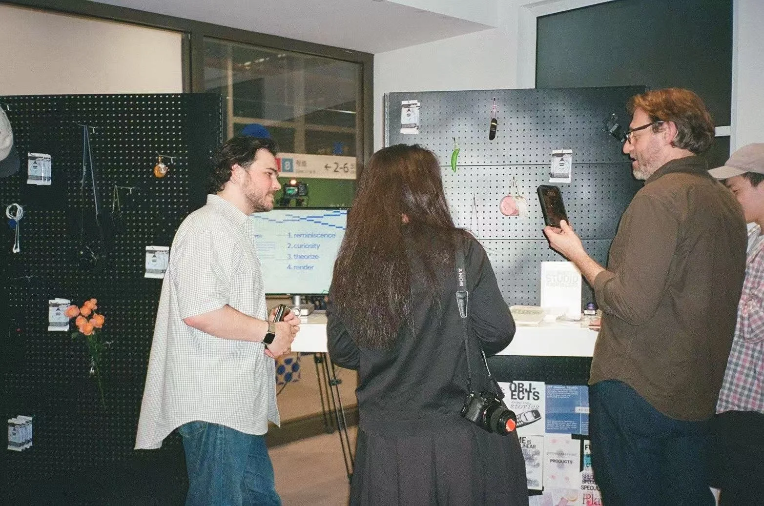 People standing and interacting at a booth with black pegboard display, hanging items, and a monitor showing a presentation, in an indoor setting.