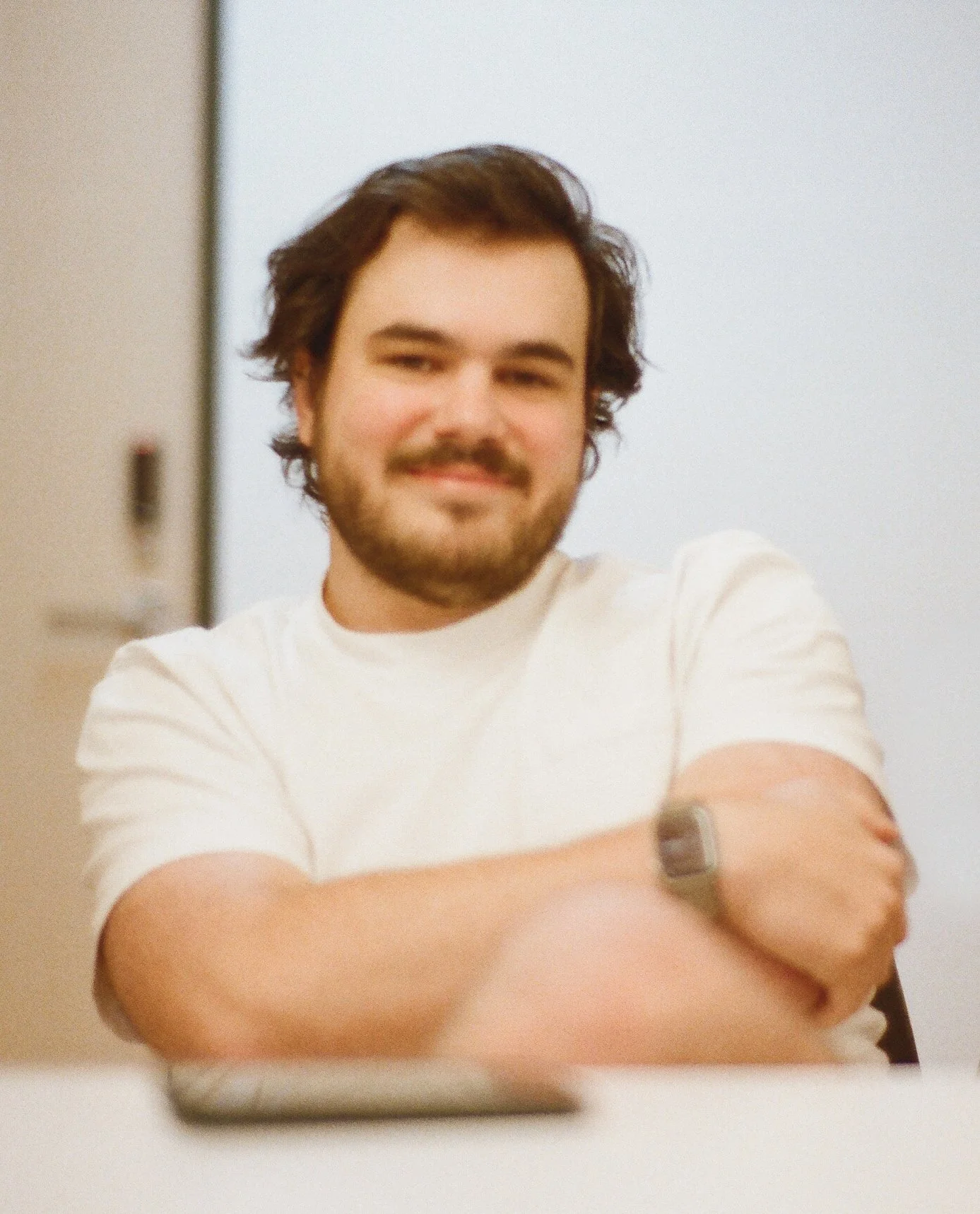 Matt Bowe - A man with dark brown hair, a beard, and a mustache, smiling with arms crossed wearing a white t-shirt and a watch, sitting indoors.
