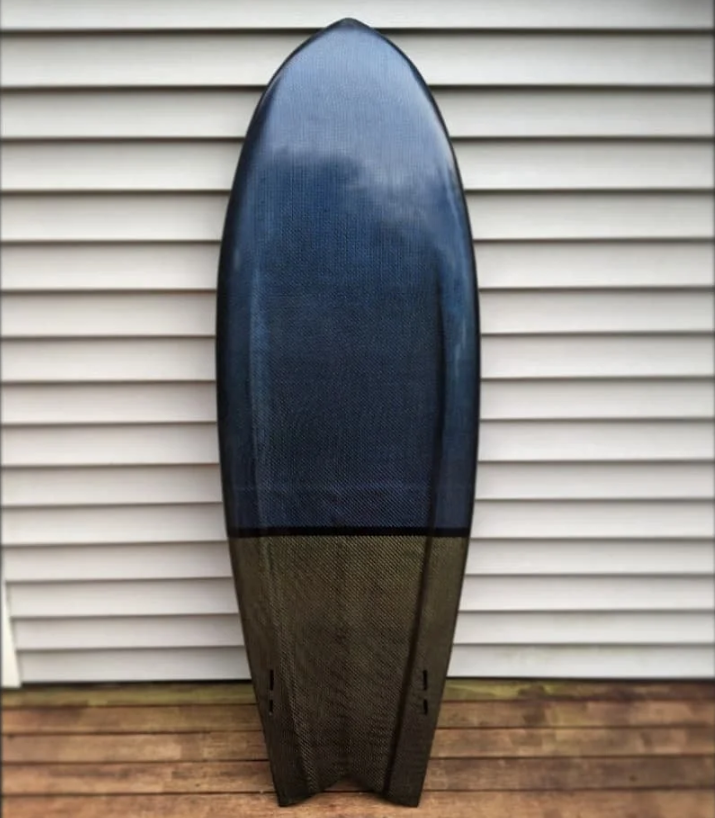 A surfboard with a dark top and a lighter bottom, standing upright against a white siding wall on a wooden deck.