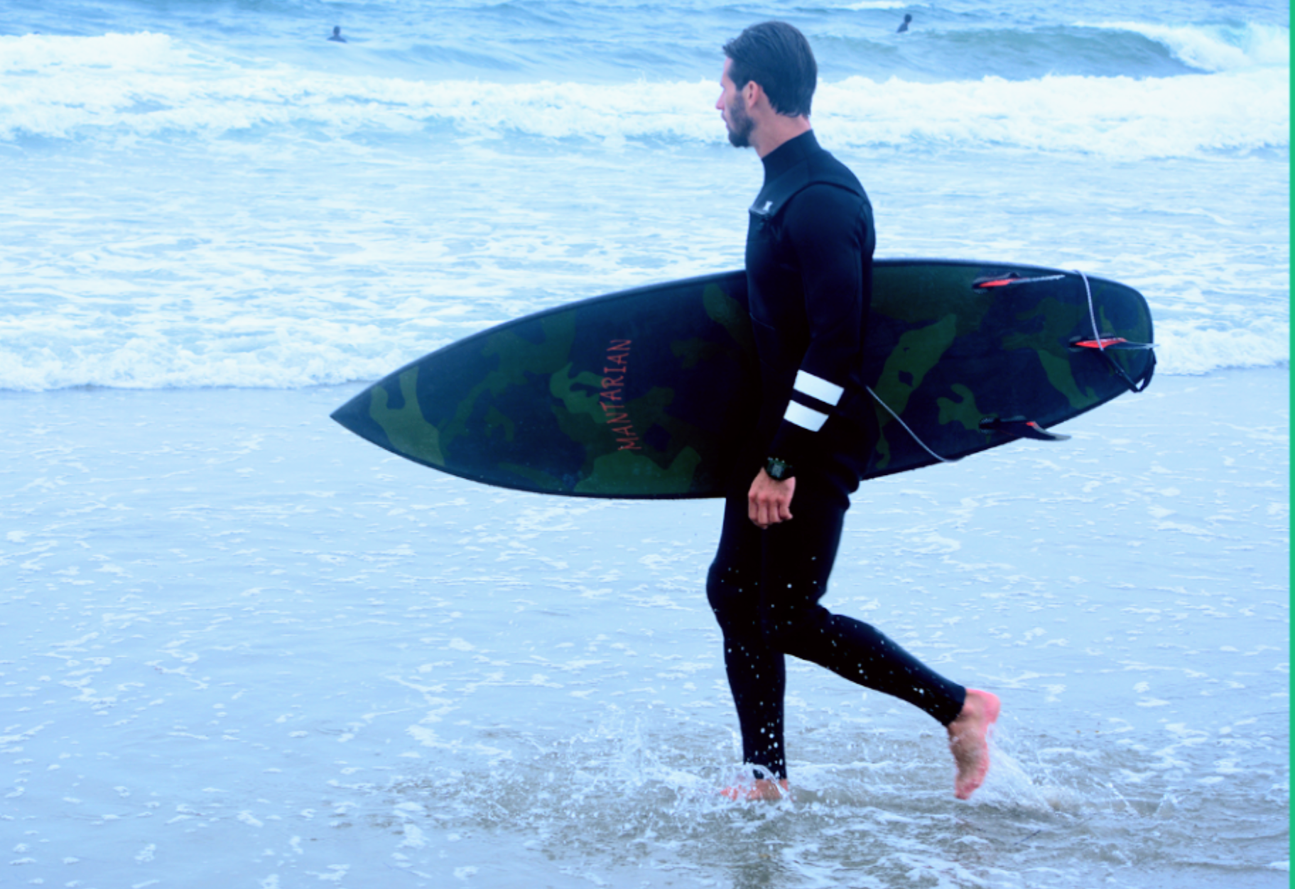 A man in a black wetsuit walking through shallow ocean water while carrying a Mantarian Designs camouflage-patterned surfboard with red and black fins.