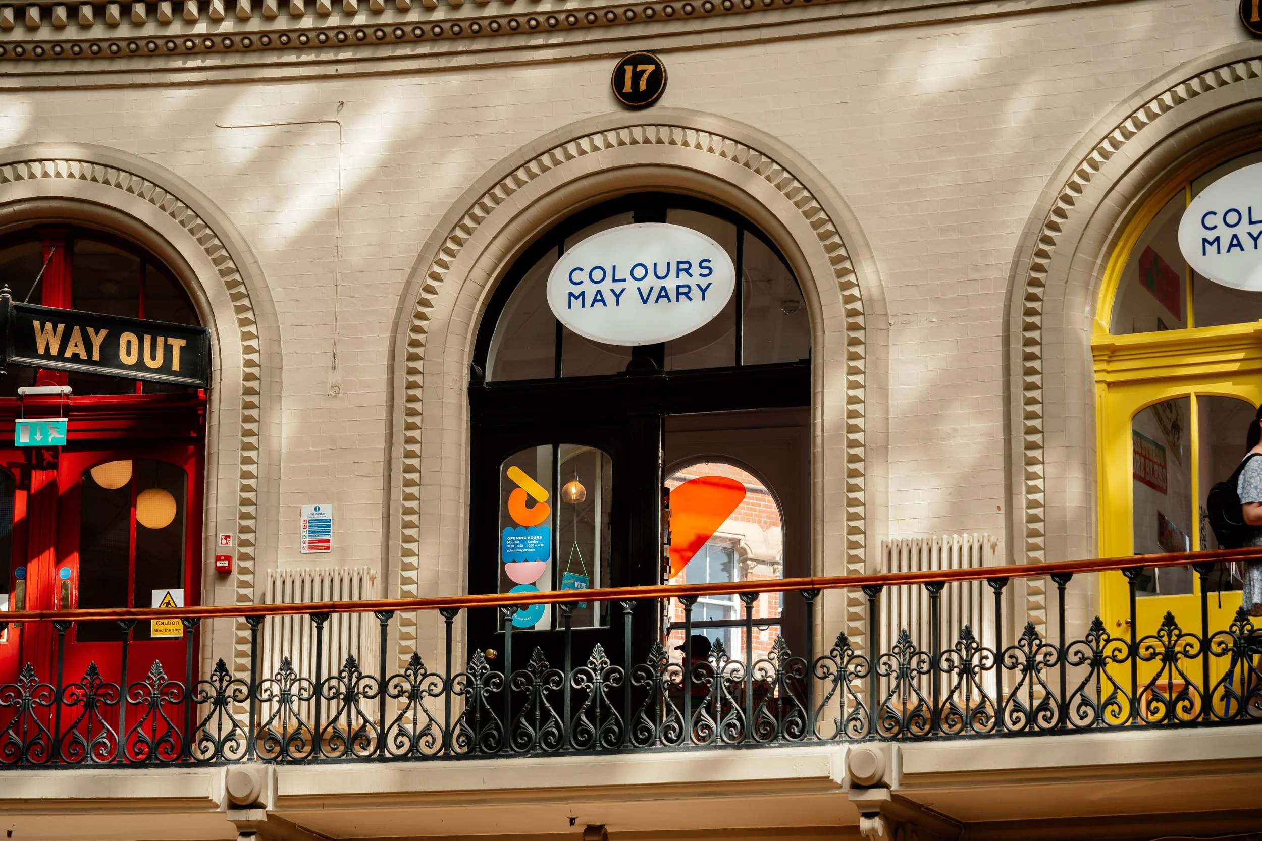 Close-up view of a storefront with a sign that says 'COLOURS MAY VARY' in an oval shape above the entrance. The building has a decorative archway with a number 17 on a black and gold plaque. Additional signage and a red door are partially visible.