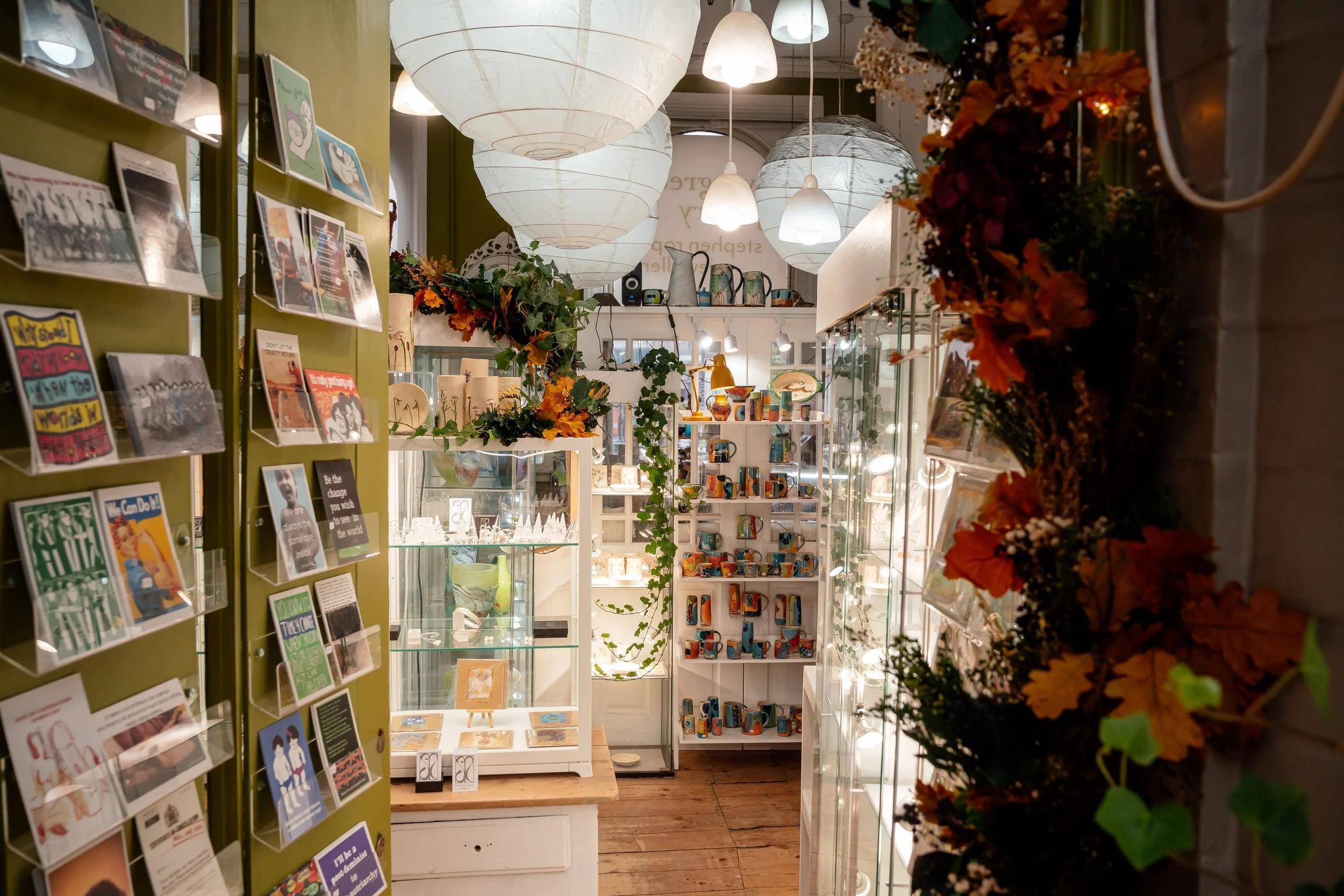 Interior of a store with hanging white paper lanterns, shelves filled with colorful mugs, and a green wall displaying postcards and artwork. Floral decorations with orange and brown leaves are on the right side.