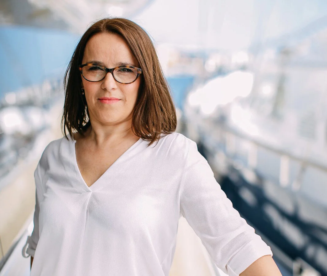 Woman with glasses wearing a white top, standing outdoors with a blurred cityscape in the background.