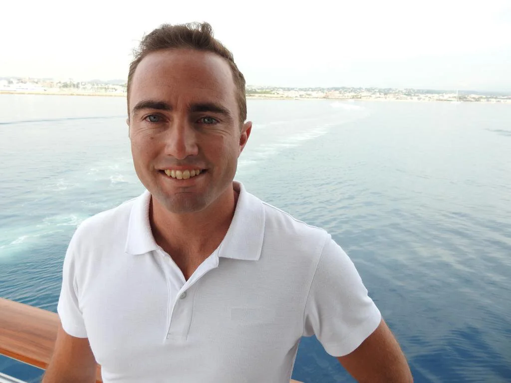 A man wearing a white polo shirt smiling on a boat, with water and a distant shoreline in the background.