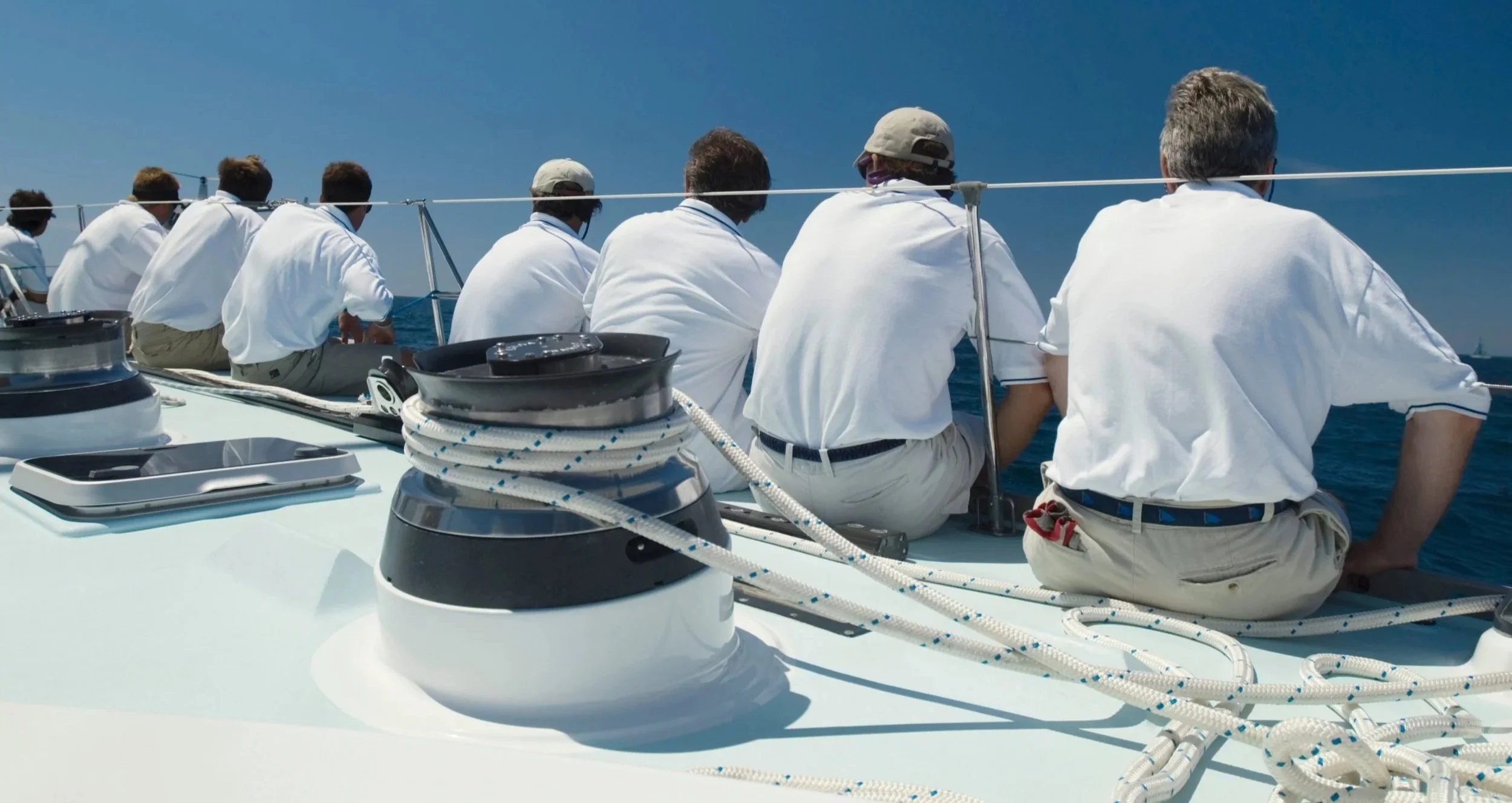 Group of people dressed in white shirts sitting on a sailboat, overlooking the ocean under a clear blue sky.