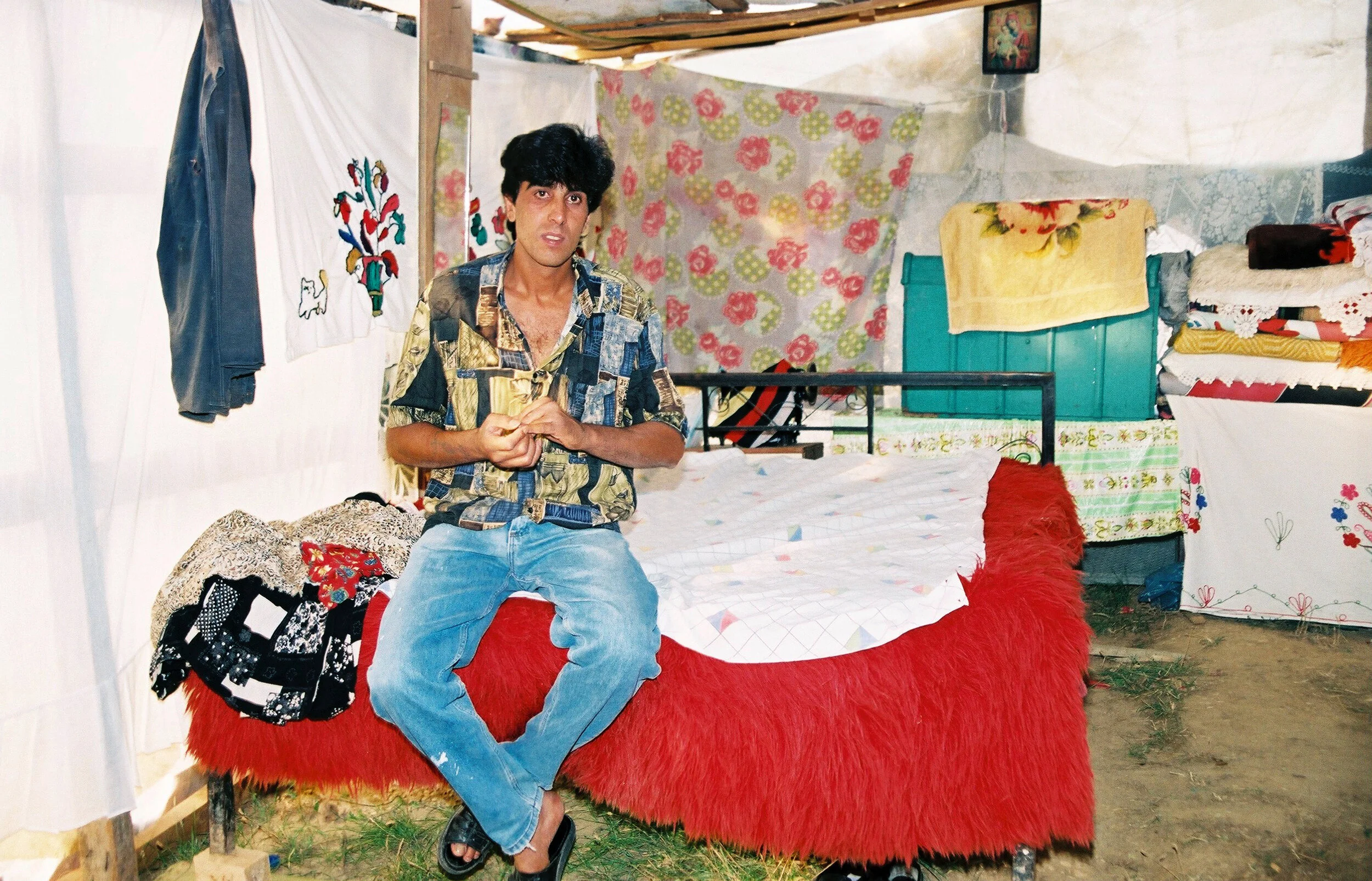 A young man with dark hair wearing a patterned shirt and jeans, sitting on a red, furry-covered bed inside a makeshift tent. The tent has various linens, towels, and cloths hanging or stacked around.