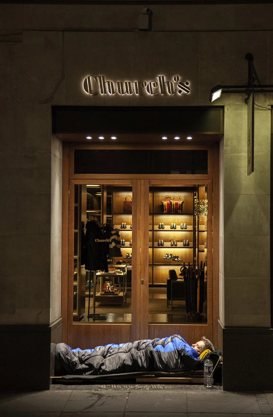 A man sleeping on a cot outside the Churchill's store at night, with the store's illuminated interior showcasing shoes, bags, and footwear on display.