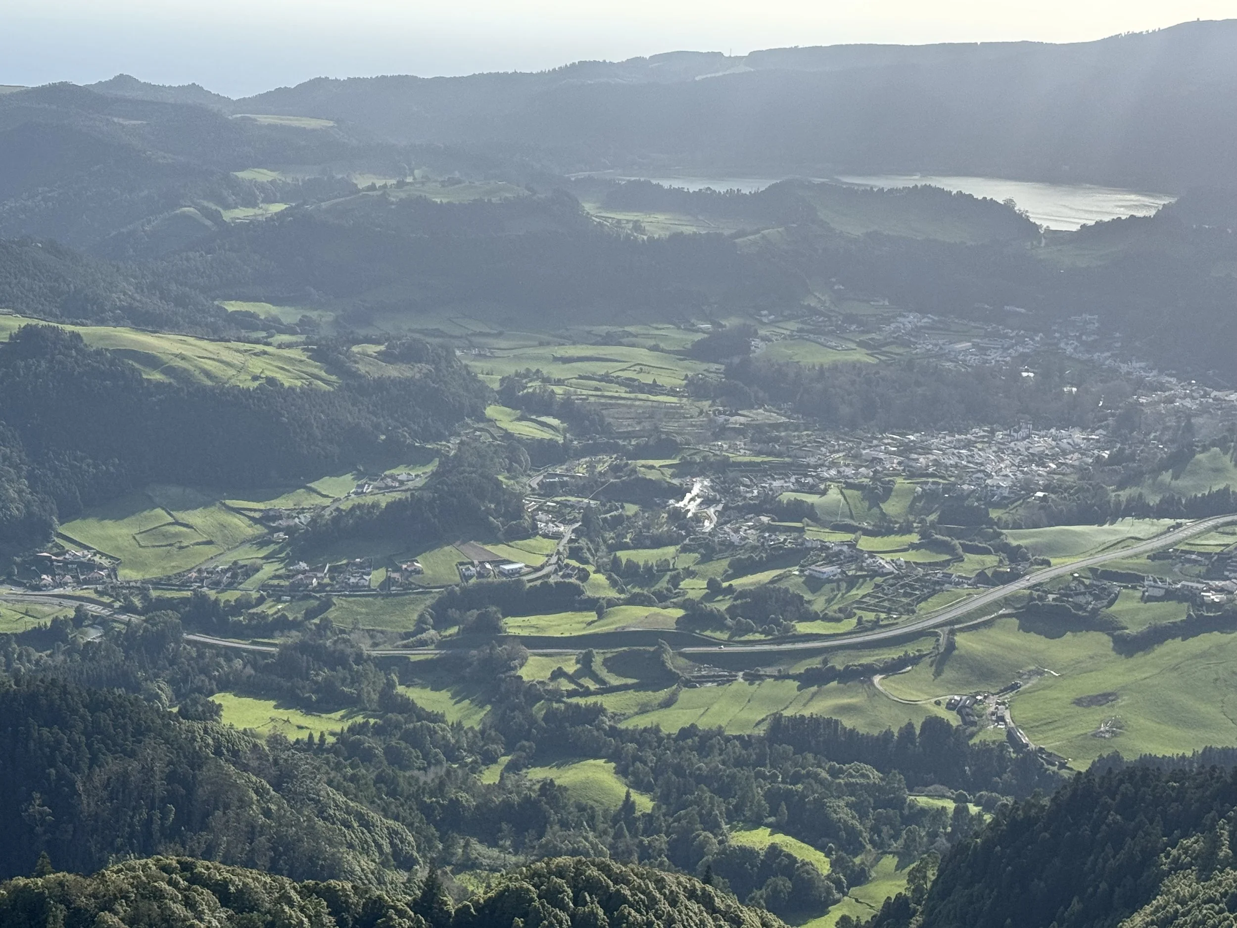 A landscape view of a green valley with small buildings and winding roads, surrounded by rolling hills and distant mountains under a clear sky.