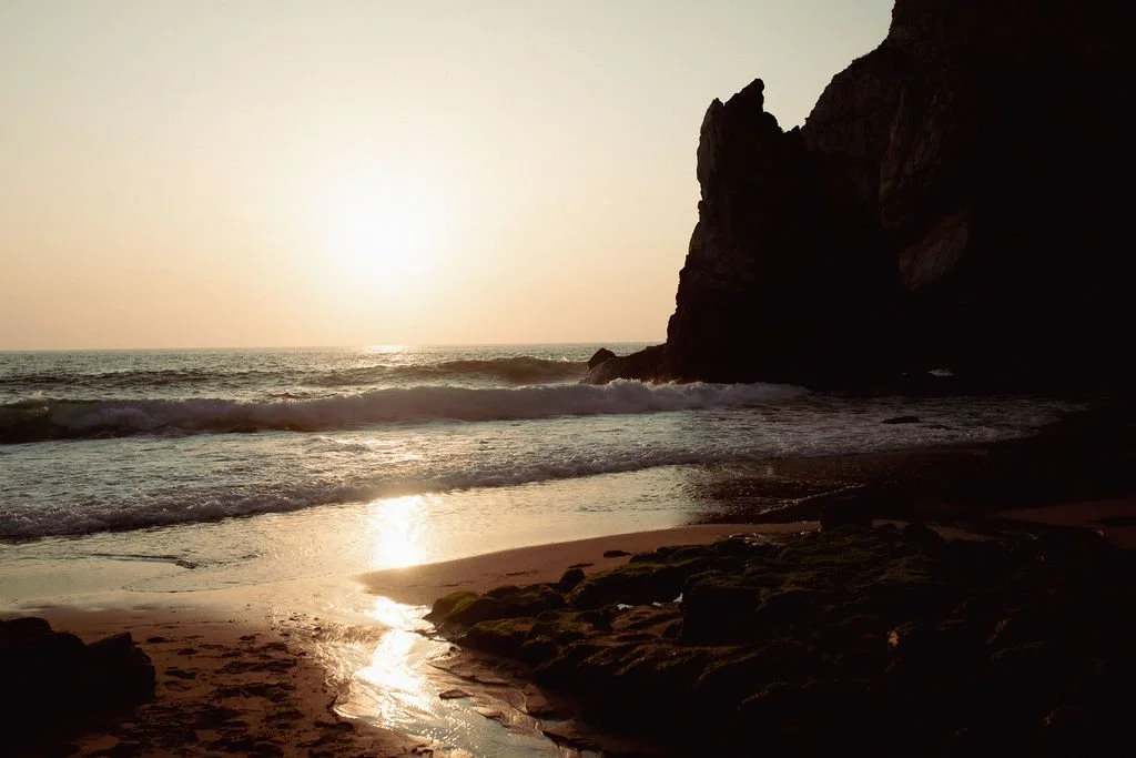 A scenic beach at sunset with the sun low on the horizon, ocean waves approaching the shore, a large dark rock formation on the right, and sandy beach foreground.