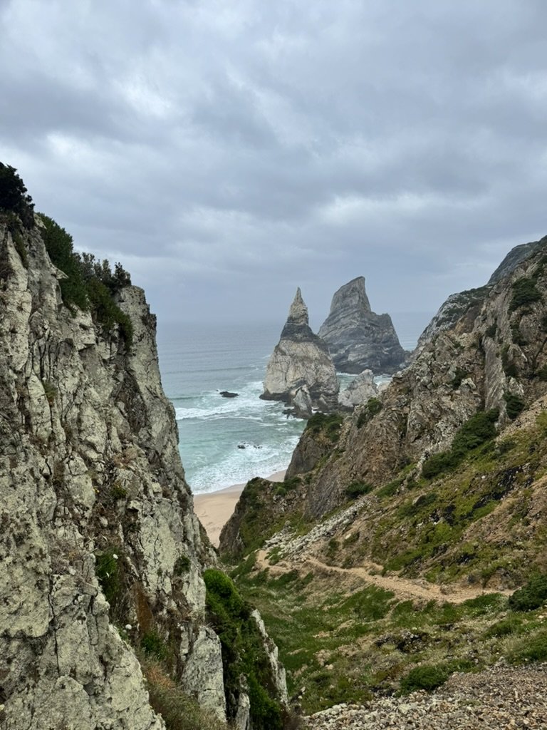 Coastal landscape with steep cliffs, rocky sea stacks, a sandy beach, and overcast sky.