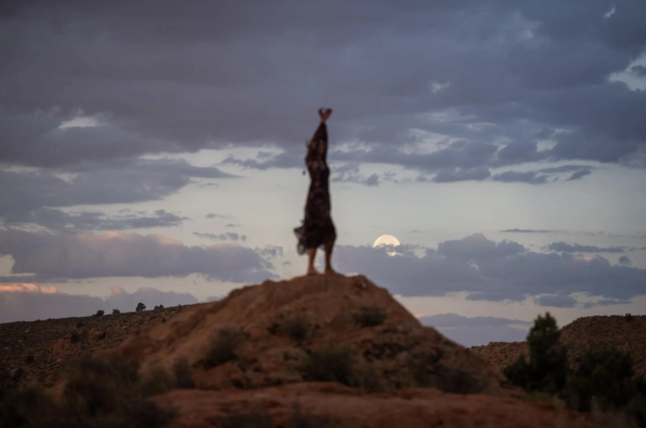 A person doing a handstand on a rock at sunset with the moon visible in the sky, over a landscape of hills and clouds.