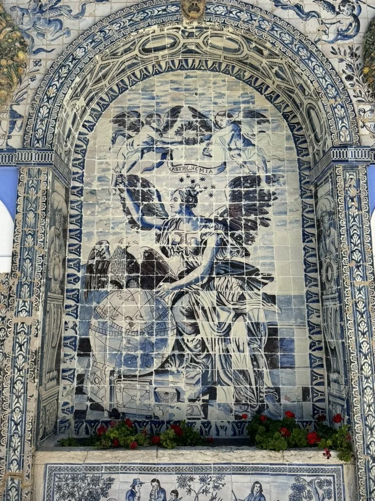 Ceramic tile mural depicting a mythological scene with a winged woman holding a staff and a globe, surrounded by ornate blue and white decorative borders.