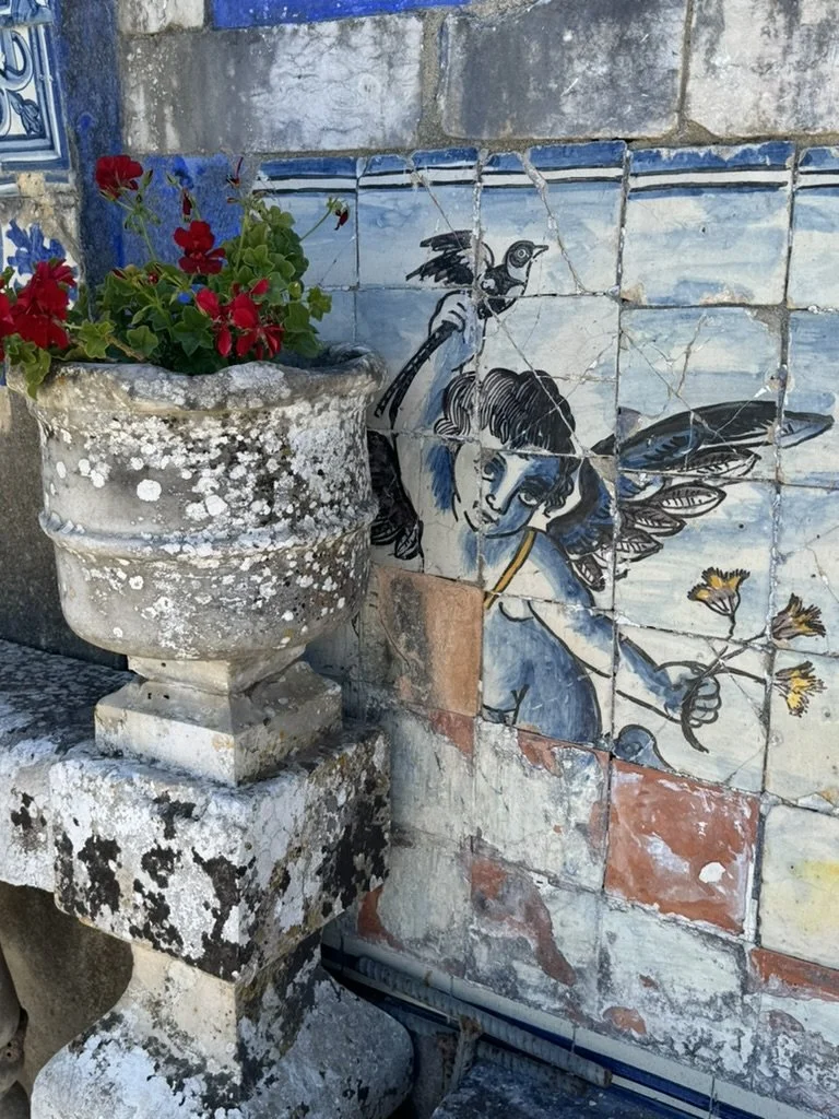 A weathered stone garden planter with red and purple flowers placed next to a tiled wall featuring a mural of a angelic figure with wings and a bird perched on its shoulder, holding a branch with yellow flowers.