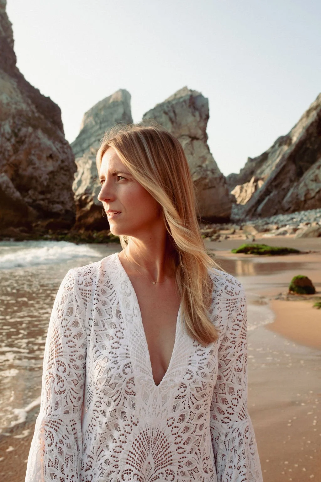 A woman with long blonde hair wearing a white lace dress standing on a beach with large rocky formations in the background, during late afternoon or early evening.