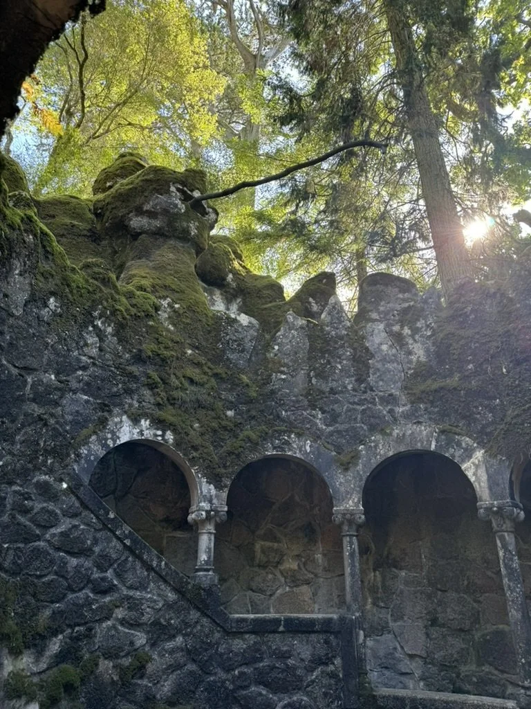 An outdoor scene of rocky terrain covered in moss with three stone arches supported by columns, surrounded by tall trees with sunlight filtering through the leaves.