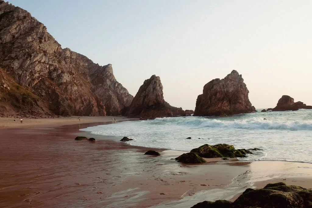 A beach with large rock formations and cliffs in the background, gentle waves washing onto the sandy shore.