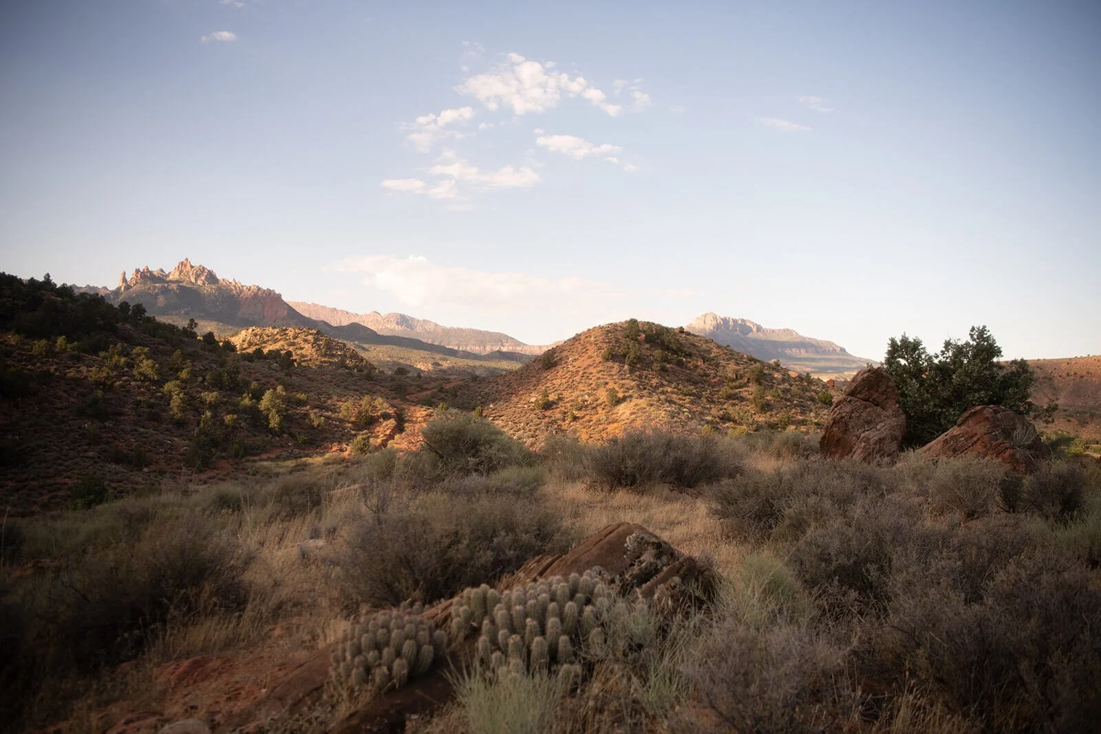 Desert landscape with rolling hills, mountain peaks in the distance, sparse vegetation, and large rocks, under a partly cloudy sky.