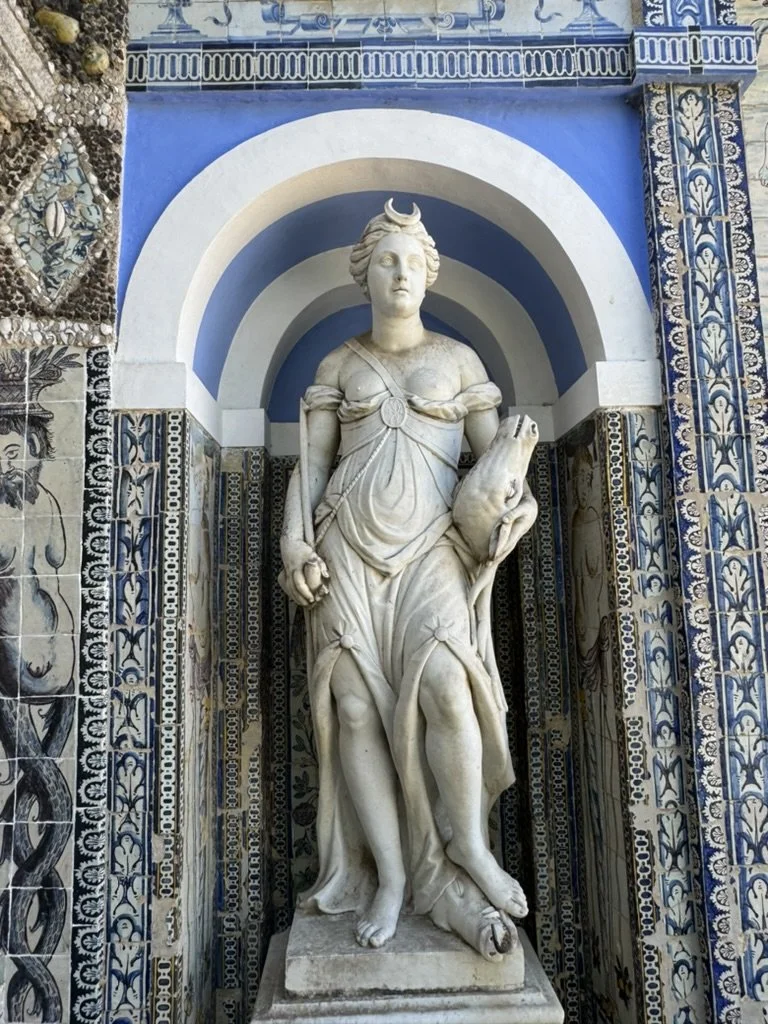 Marble sculpture of a woman with a crescent moon on her head, holding a frog, set within an arched alcove decorated with blue and white mosaic tiles.