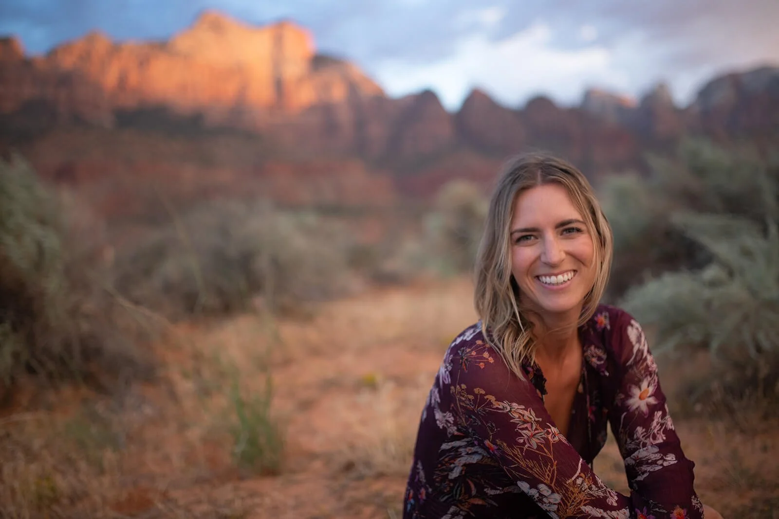 A woman with blonde hair smiling, wearing a maroon floral blouse, sitting in a desert landscape with mountains in the background during sunset.