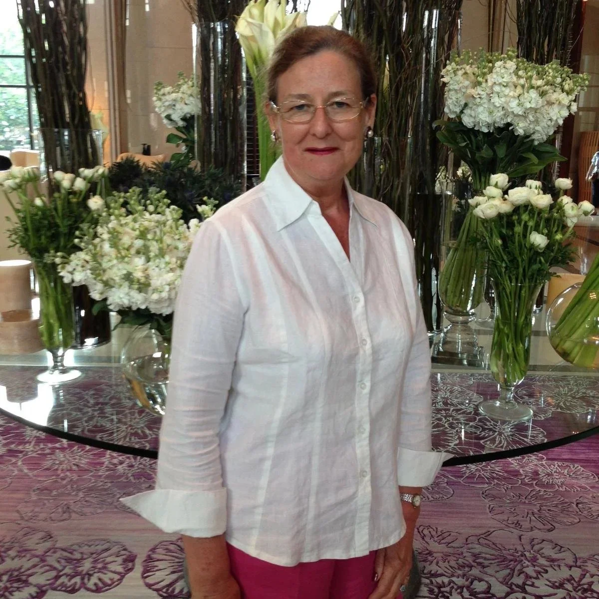 An older woman with glasses and earrings standing in front of a floral arrangement with white flowers in vases, in a well-lit room with large windows and curtains.