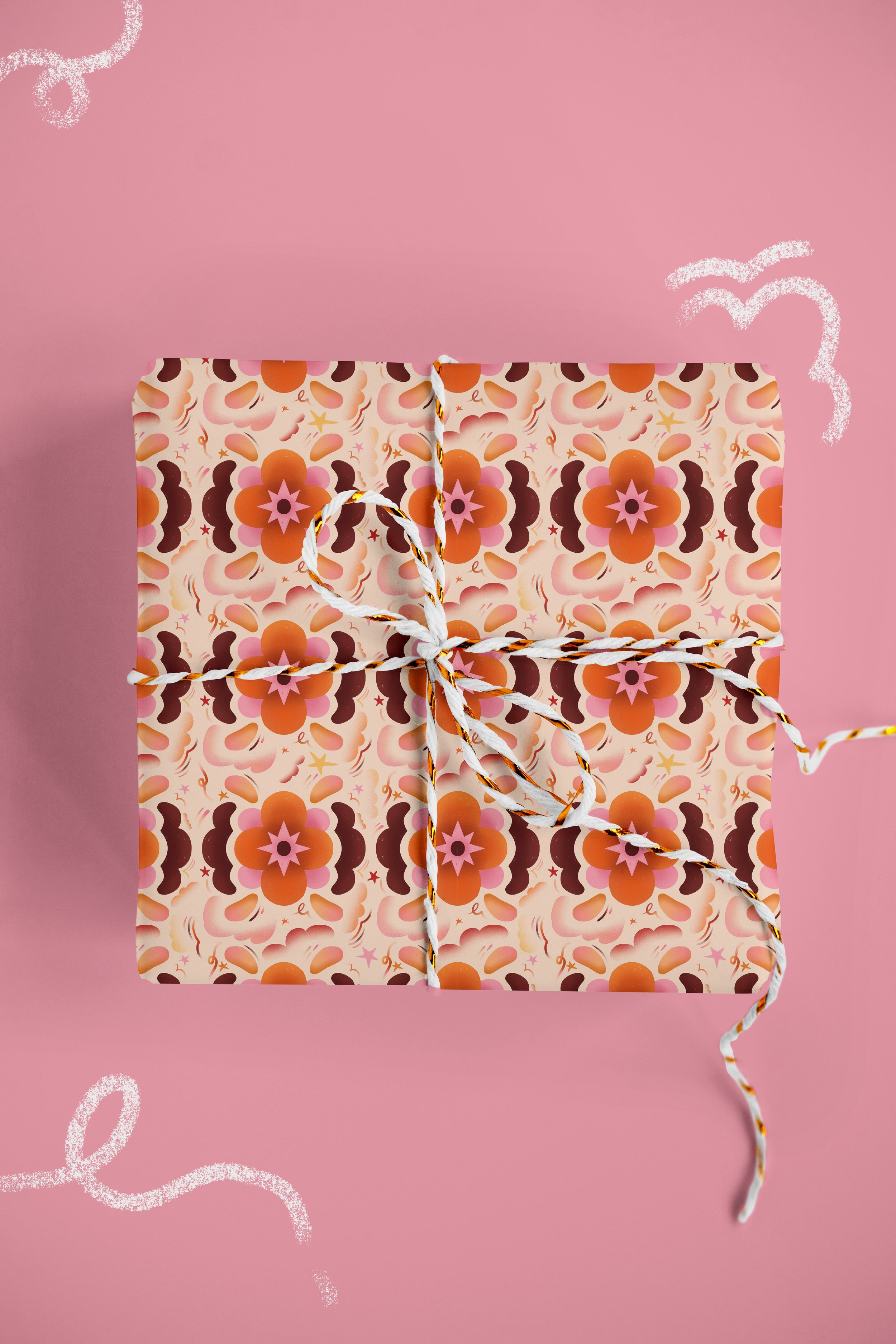 A gift wrapped in floral patterned paper with a marroon, pink, and orange color scheme, tied with a white and orange twisted string, placed on a pink background with white chalk-like squiggles.