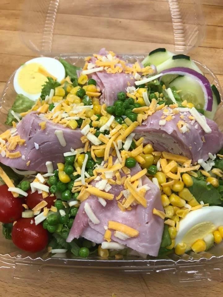 A salad with cherry tomatoes, hard-boiled egg slices, cucumber slices, ham, green peas, corn, shredded cheese, and lettuce in a clear plastic container on a wooden surface.