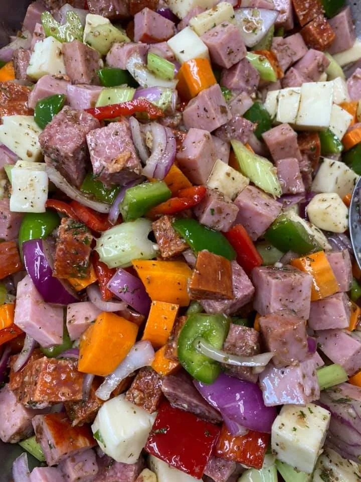 Close-up of a colorful chopped vegetable and meat salad with diced ham, green, red, and orange bell peppers, onions, and small pieces of cooked meat or sausage.