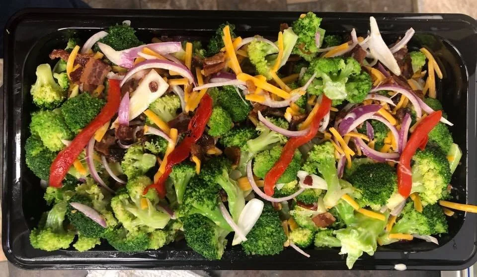 Mixed broccoli, red bell peppers, shredded cheddar cheese, shredded lettuce, and bacon bits in a takeout container.