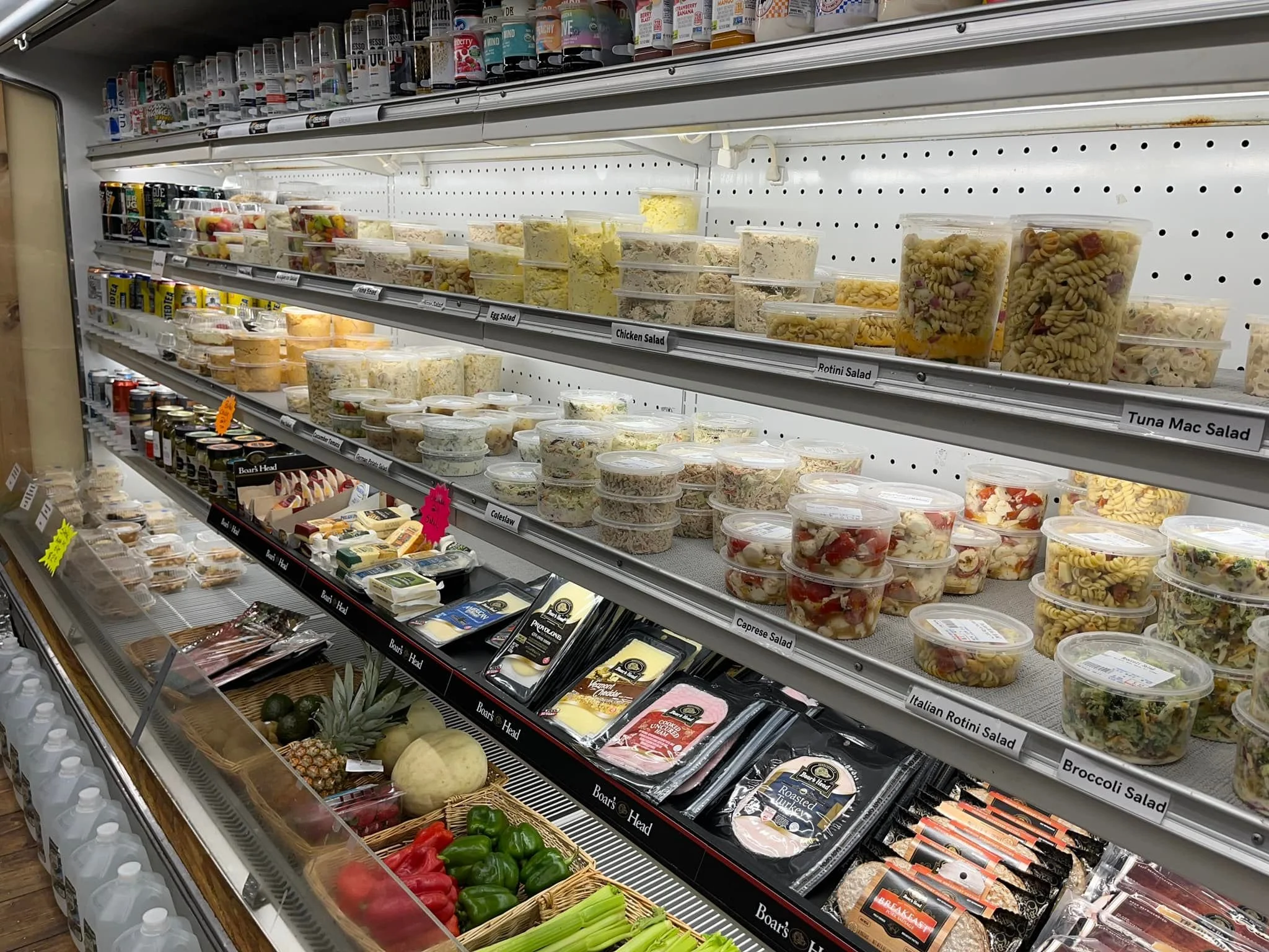 Refrigerated grocery shelf displaying prepackaged salads including egg salad, chicken salad, rotini salad, tuna mac salad, caprese salad, Italian rotini salad, and broccoli salad, with fresh fruits and vegetables nearby.