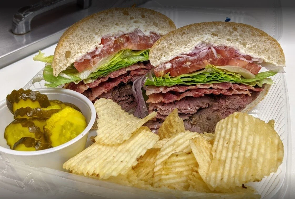 A pastrami sandwich with lettuce, tomato, and onions on a sesame bun, served with potato chips and a cup of pickles.