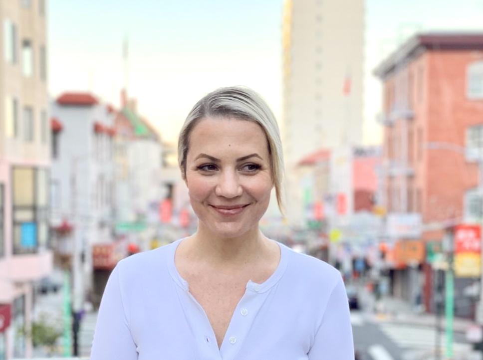 Julianne Newton, the owner of San Francisco colonics, woman with short blonde hair wearing a light blue shirt standing on a city street during daytime with colorful buildings and blurred background.