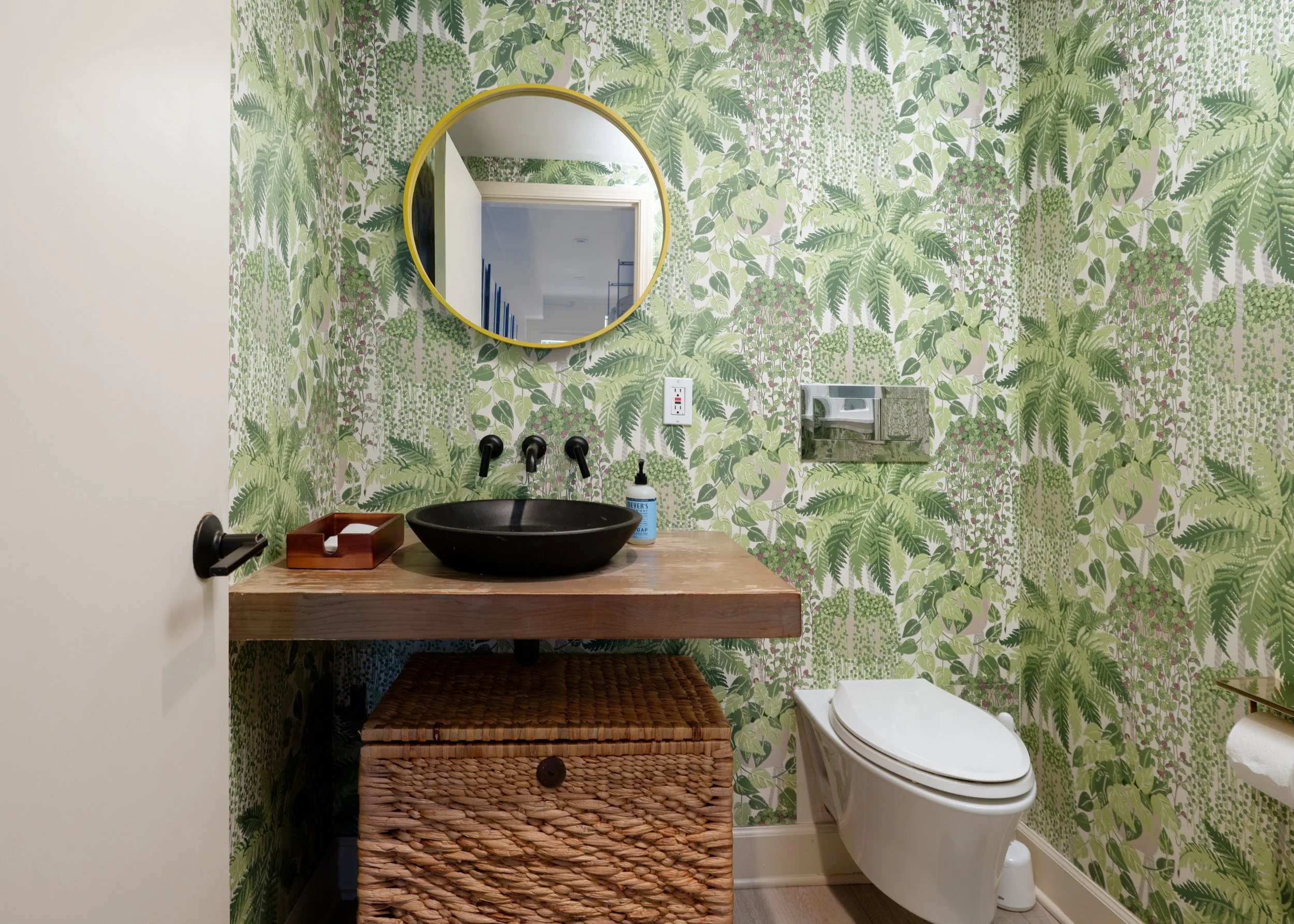 Stylish bathroom with botanical wallpaper, black vessel sink, round gold-framed mirror, wooden vanity, and white toilet