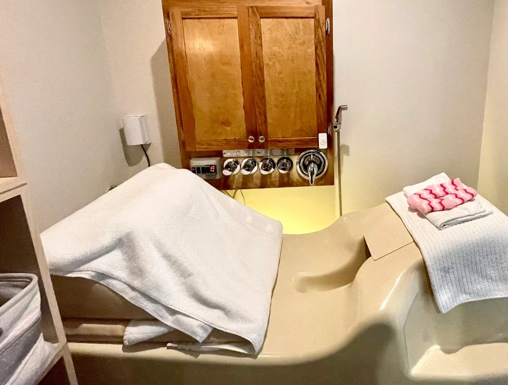 A hospital room with a beige bathtub and a white towel, a folded towel with a pink and white flower pattern on top, and a wooden cabinet in the background.
