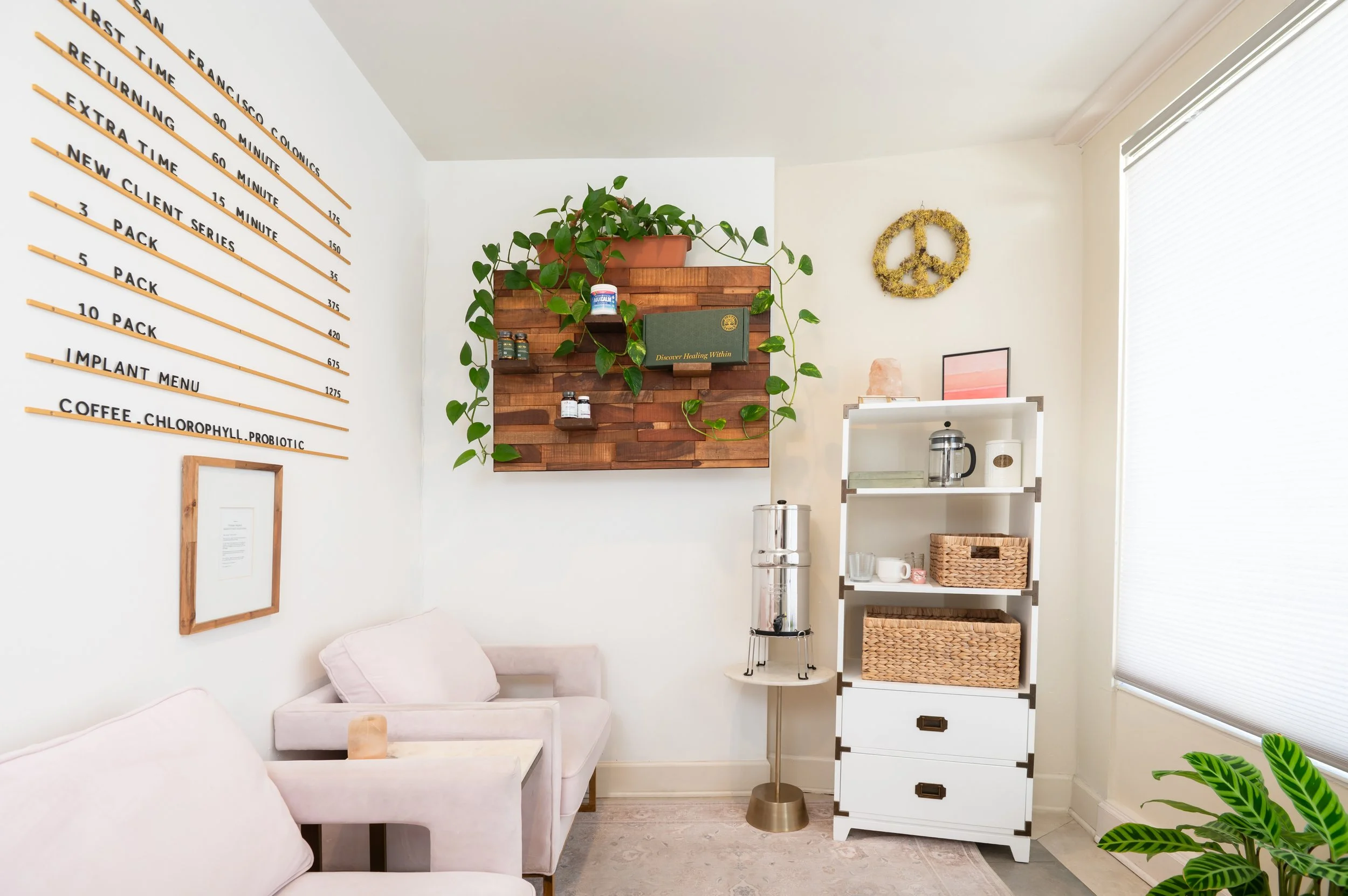 Bright studio waiting room with a treatment menu wall, plants, wooden display, tea station, cozy armchairs, and large window blinds for privacy