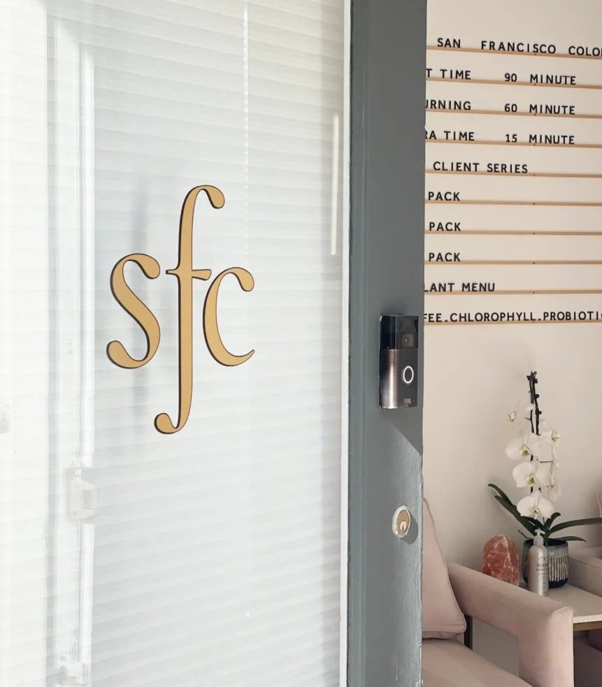Glass door with the initials 'SFC' in gold lettering, next to an interior wall with a menu board, a small table with a vase of white orchids, and seating.