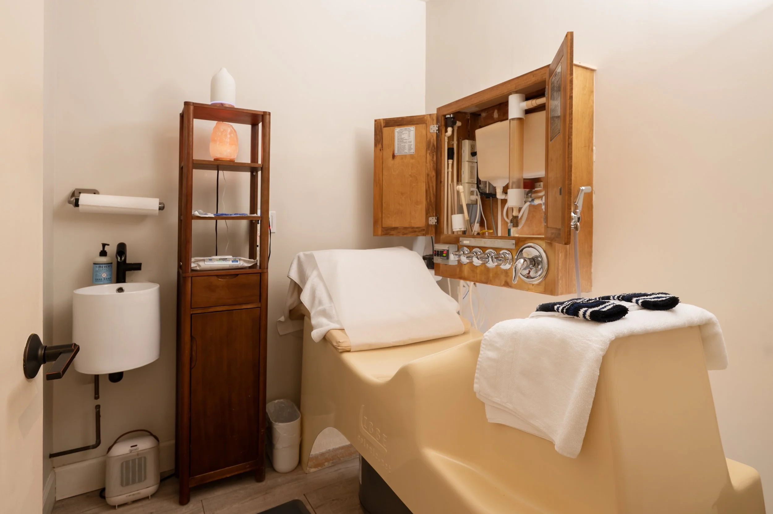Clean col;onic treatment room with a padded exam table, folded towels, sink, wooden storage cabinet, with the LIBBE equipment open.