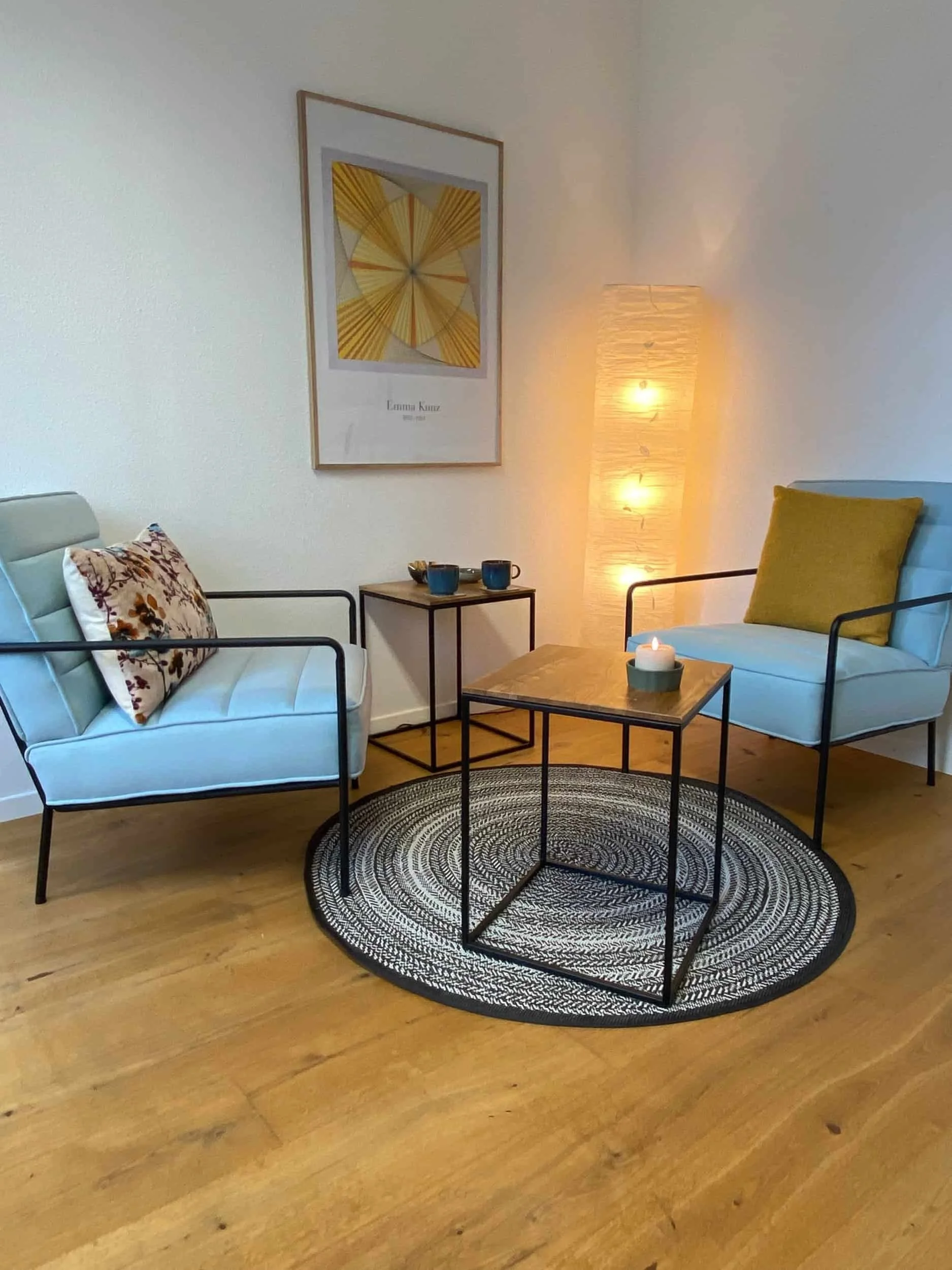 Gemütliches Wohnzimmer mit zwei weißen Sofas, einem kleinen schwarzen Beistelltisch, einem runden schwarzen und weißen Teppich, einem Holz-Tisch mit Kerze, einem breiten Standlampen, einer Wandkunst mit gelben und braunen Mustern und vier Bier- oder 