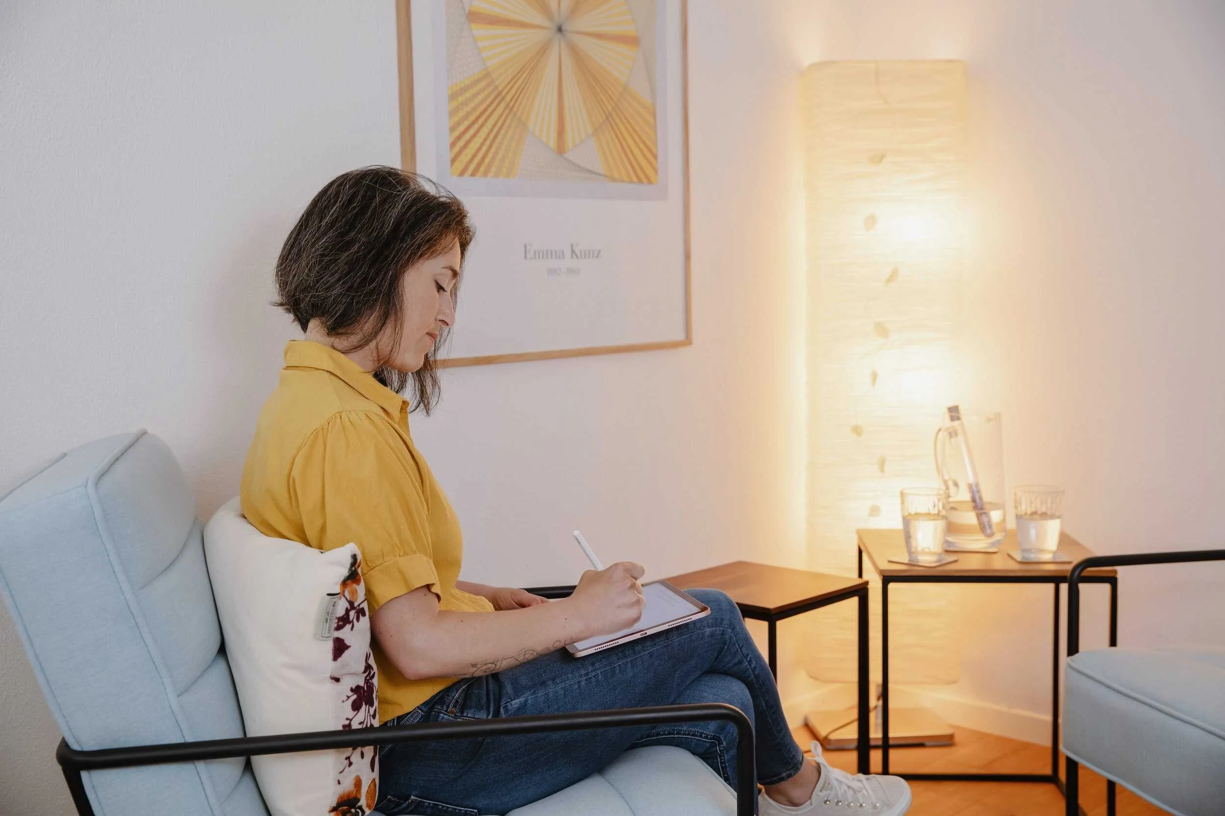 Frau sitzt auf einem Sofa, schreibt auf einem Tablet, im Hintergrund ein modernes Zimmer mit Wandbild und einem Bodenleuchte, Getränke auf einem kleinen Tisch.