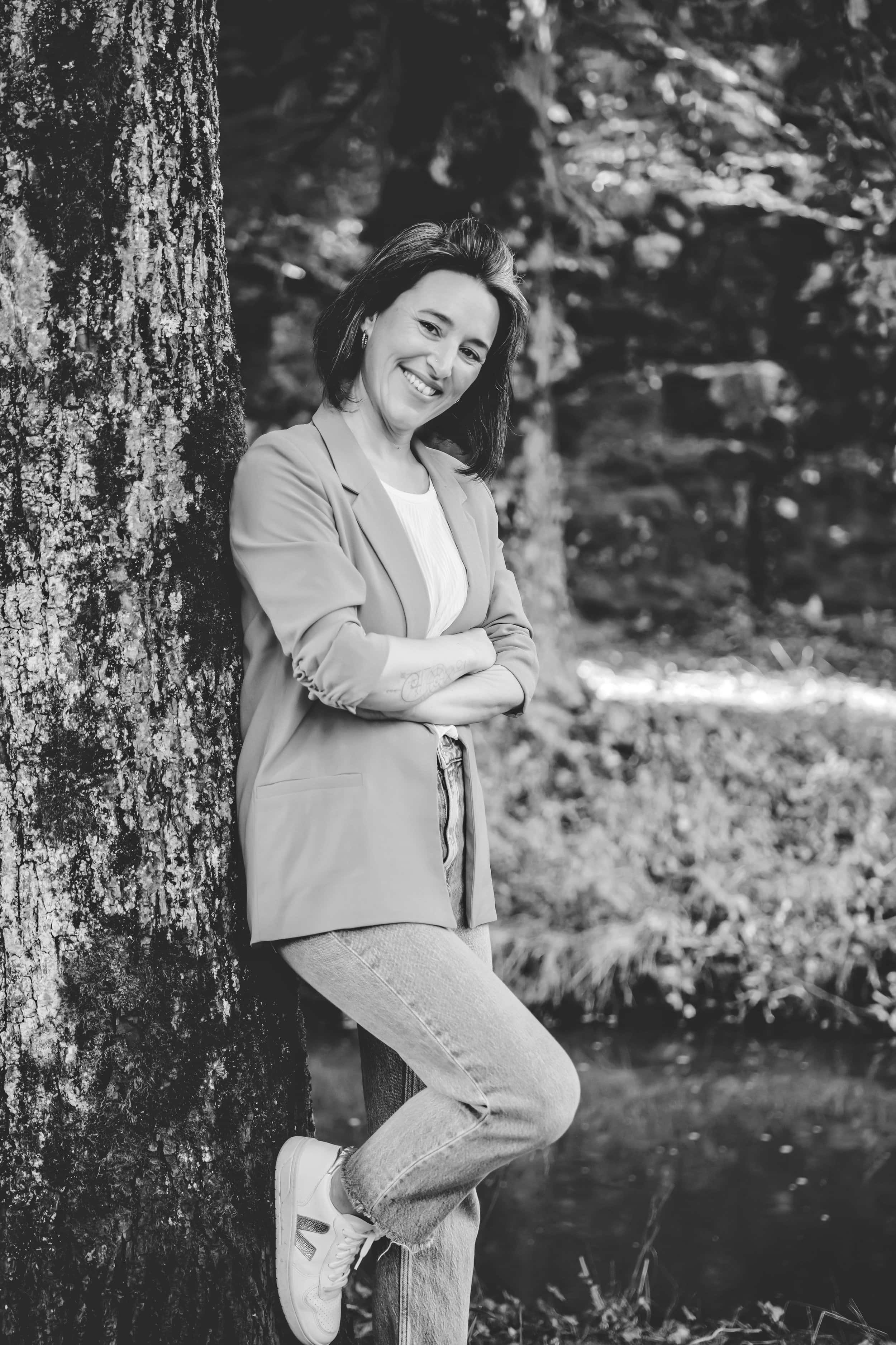 Eine lachende Frau lehnt an einem Baum im Freien, umgeben von Natur. Schwarz-weiße Aufnahme.